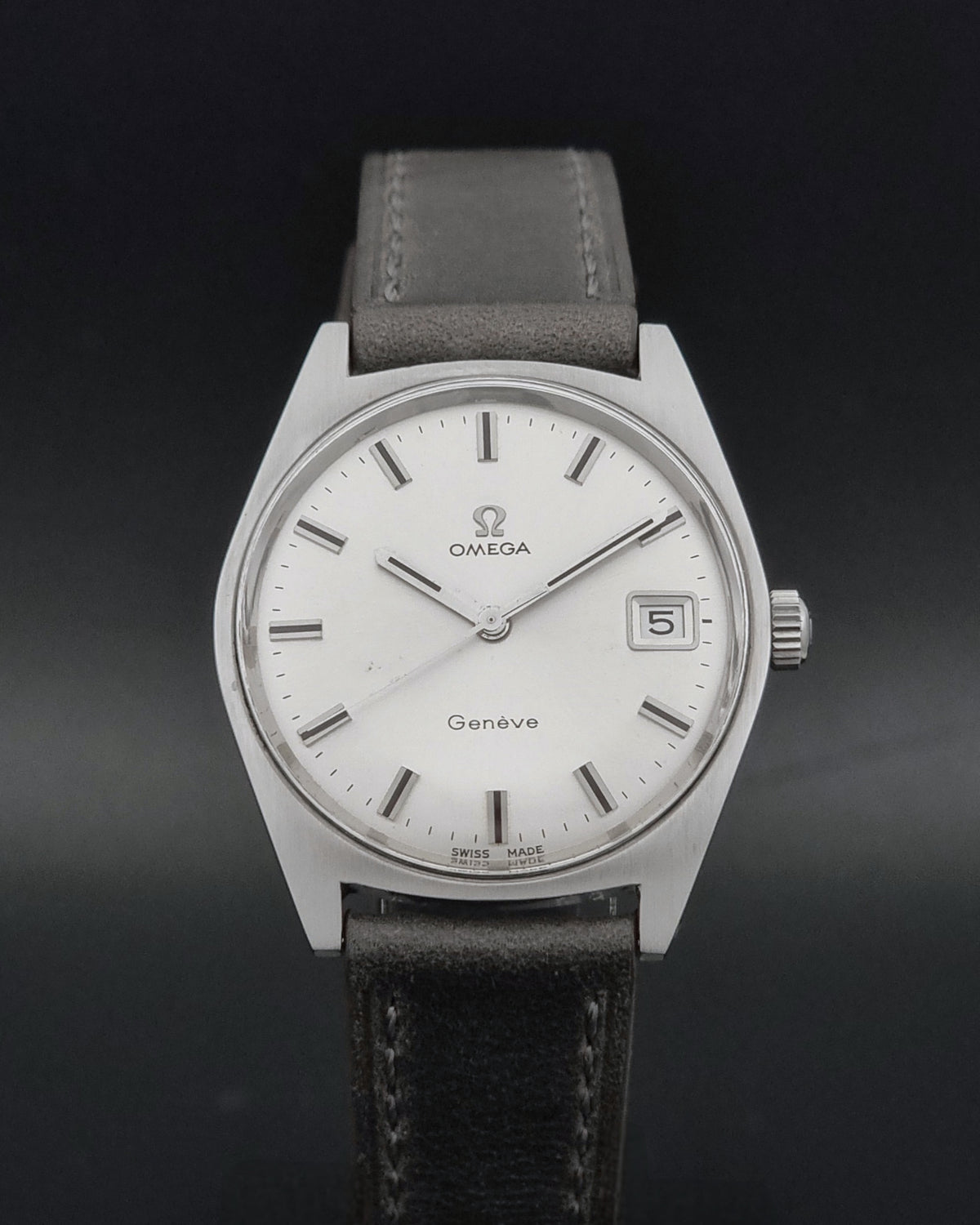 Omega Geneve wristwatch with a silver stainless steel case, white dial, and a dark gray leather strap, shown against a dark background.