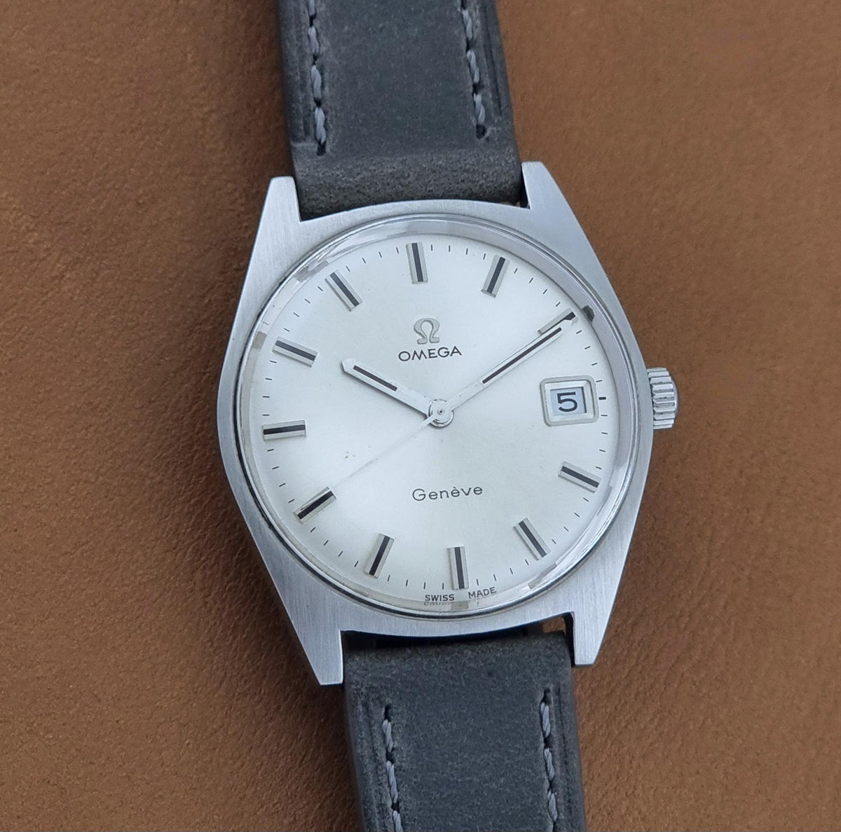 A vintage Omega Geneve wristwatch with a silver-toned metal case, white dial, and dark gray leather strap, shown against a brown textured background.