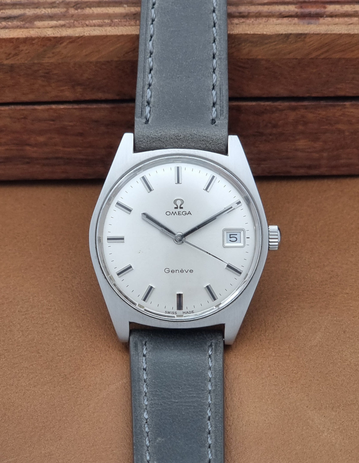 Vintage Omega Geneve 136.041 watch with a silver-toned oval case, white dial, and a gray leather strap, displayed on a brown leather surface with a wooden background.