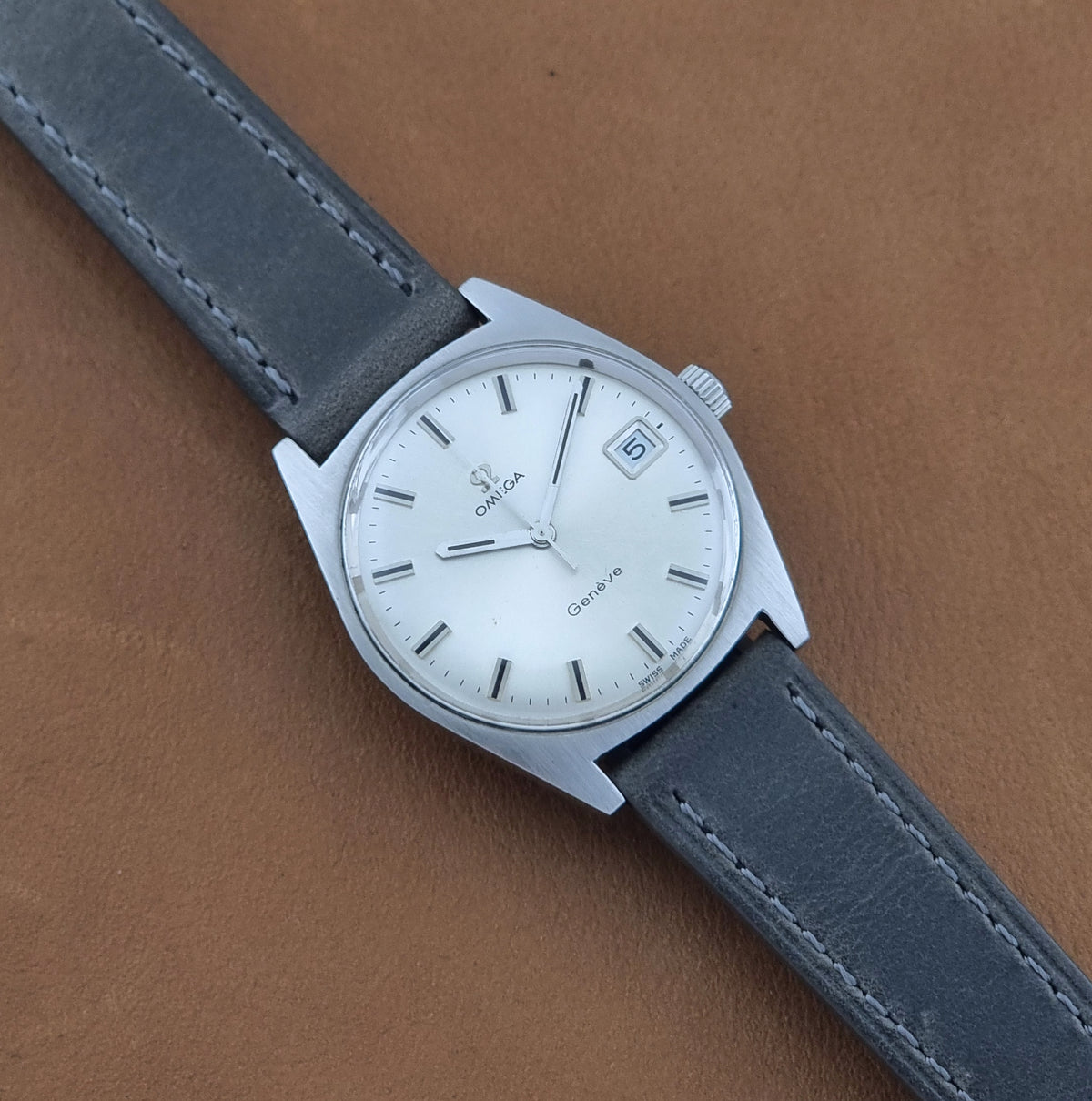 Vintage Omega Geneve 136.041 watch with a silver-toned case, white dial, and a dark gray leather strap, laid diagonally on a brown textured surface.