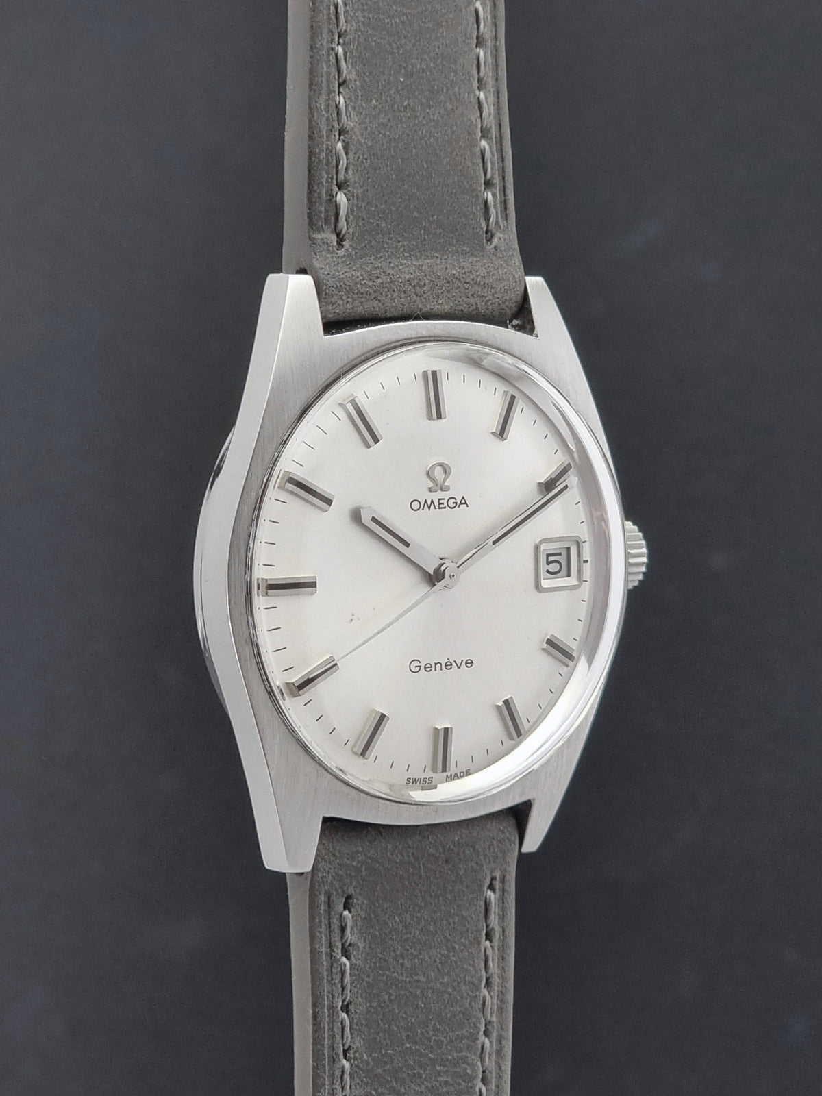 An Omega Geneve wristwatch with a silver-tone oval case, white dial, and a gray leather strap, shown against a dark gray background.