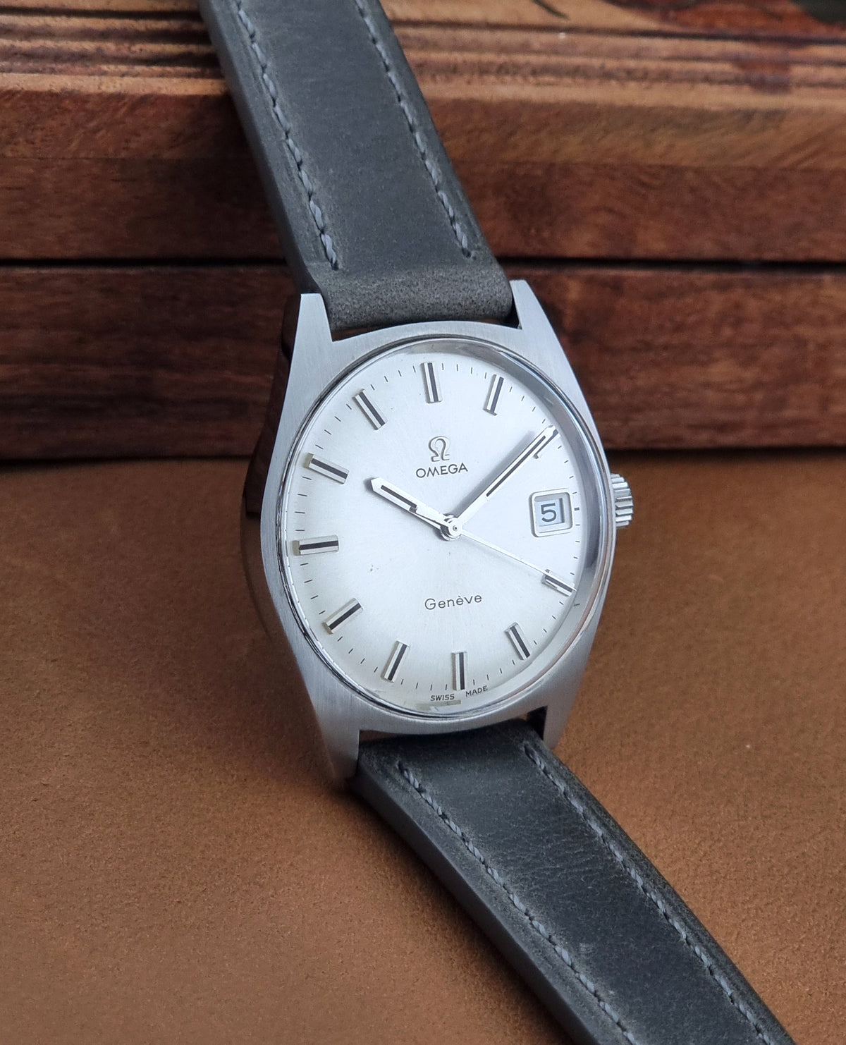 An Omega Geneve wristwatch with a silver-tone stainless steel case, white dial, and dark gray leather strap, resting on a brown leather surface with a wooden background.