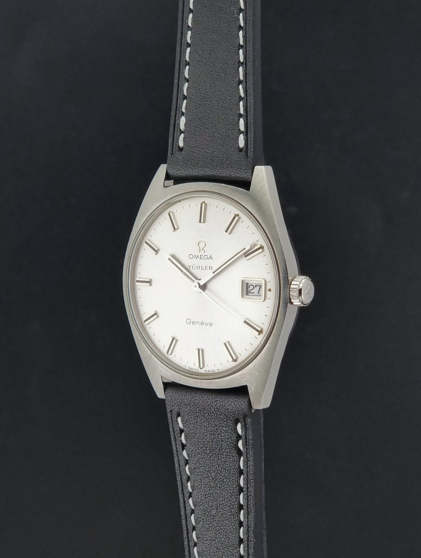 Vintage Omega watch Geneve 136.041 Co-signed Türler with a stainless steel case,  and a black leather strap with white stitching, shown on a gray background crown profile view