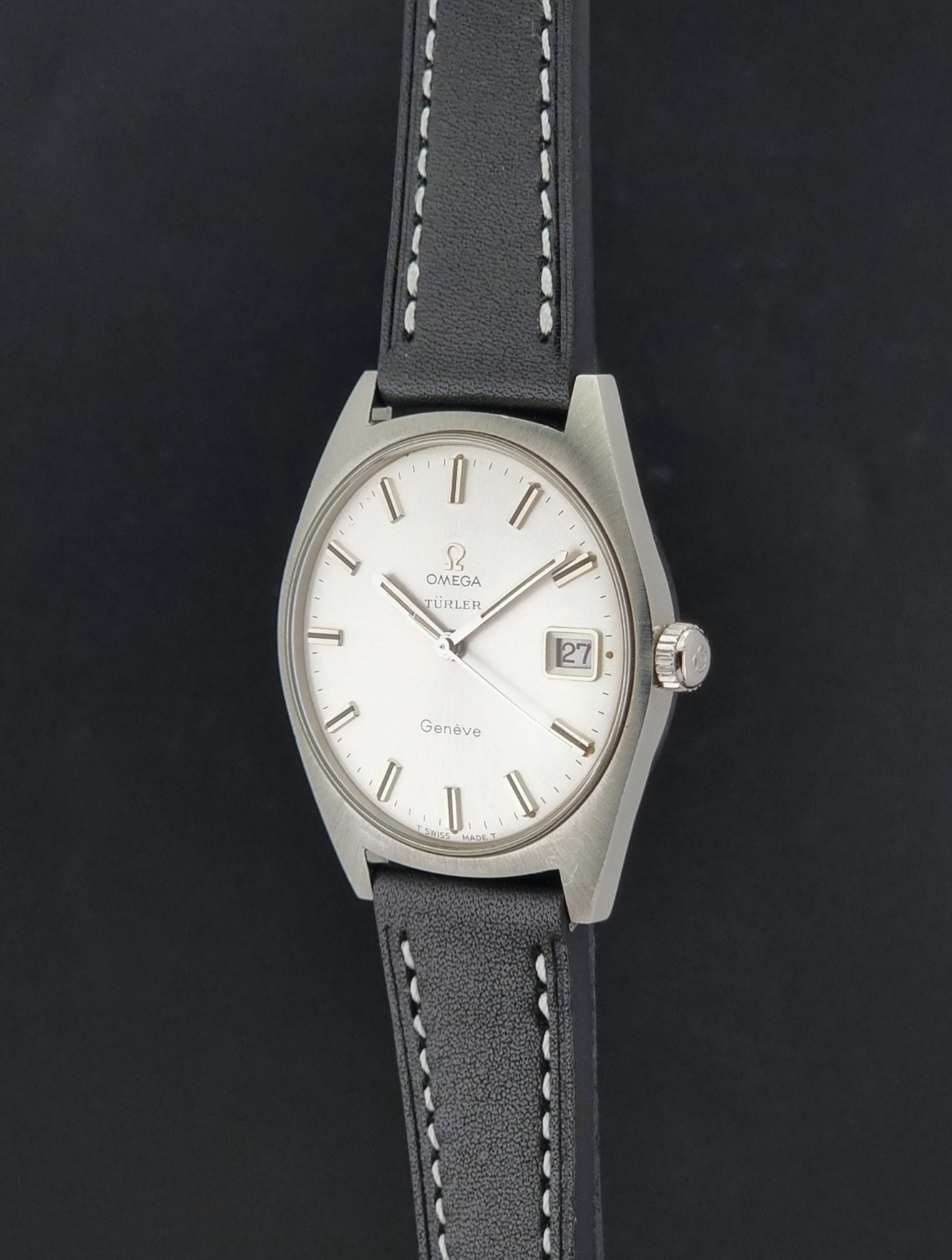 Vintage Omega watch Geneve 136.041 Co-signed Türler with a stainless steel case,  and a black leather strap with white stitching, shown on a gray background crown profile view