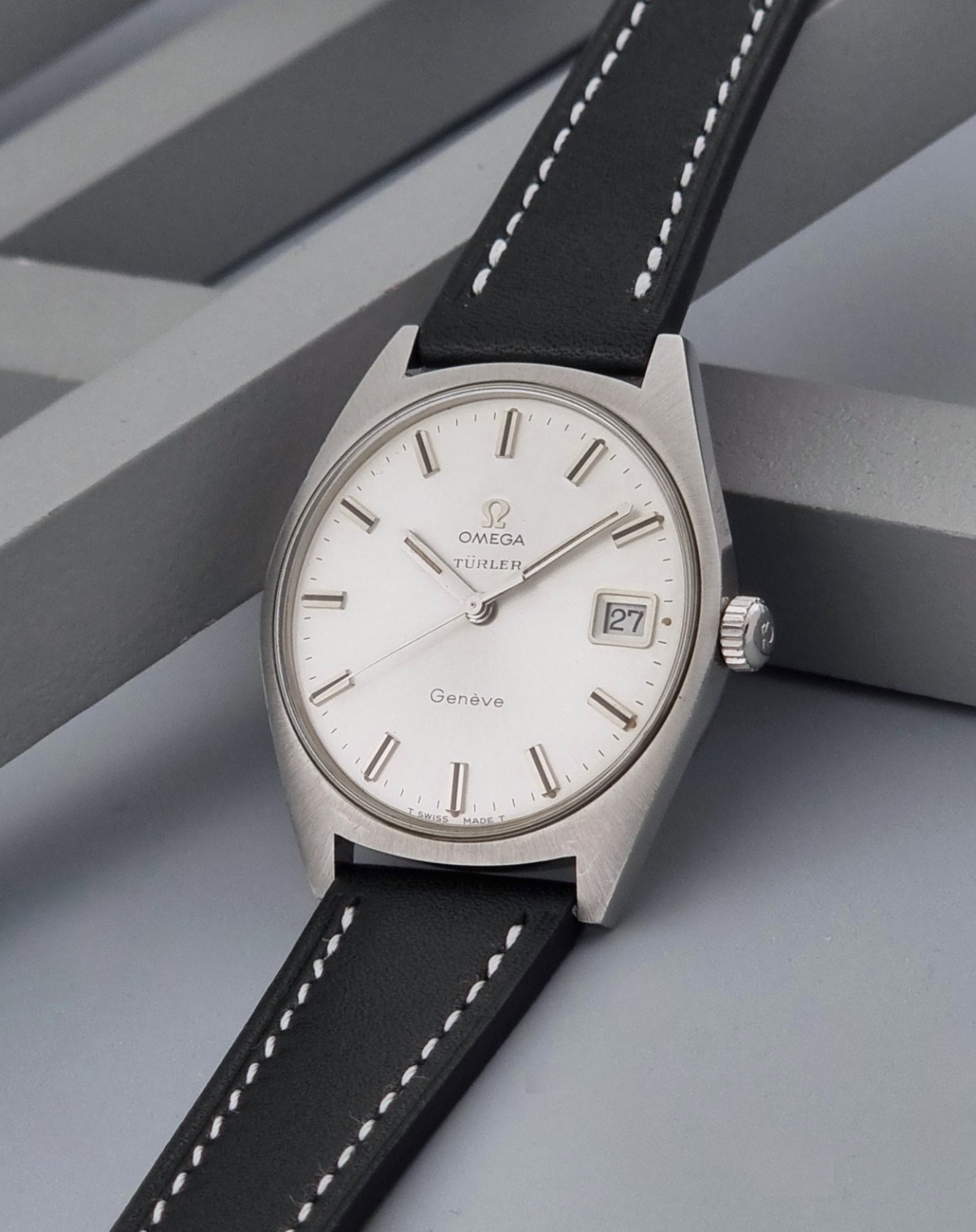Vintage Omega watch Geneve 136.041 Co-signed Türler with a stainless steel case,  and a black leather strap with white stitching, shown on a gray background crown view