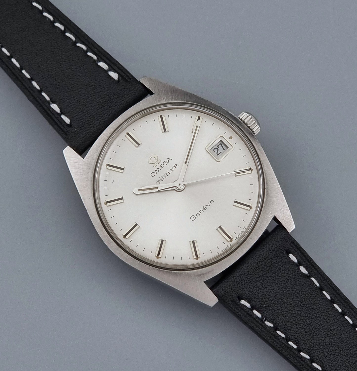 Vintage Omega watch Geneve 136.041 Co-signed Türler with a stainless steel case,  and a black leather strap with white stitching, shown on a gray background close up dial view