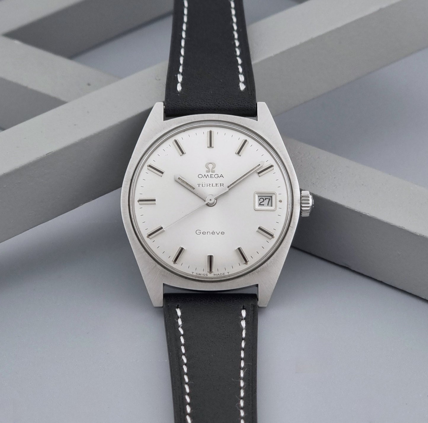 Vintage Omega watch Geneve 136.041 Co-signed Türler with a stainless steel case,  and a black leather strap with white stitching, shown on a gray background dial view