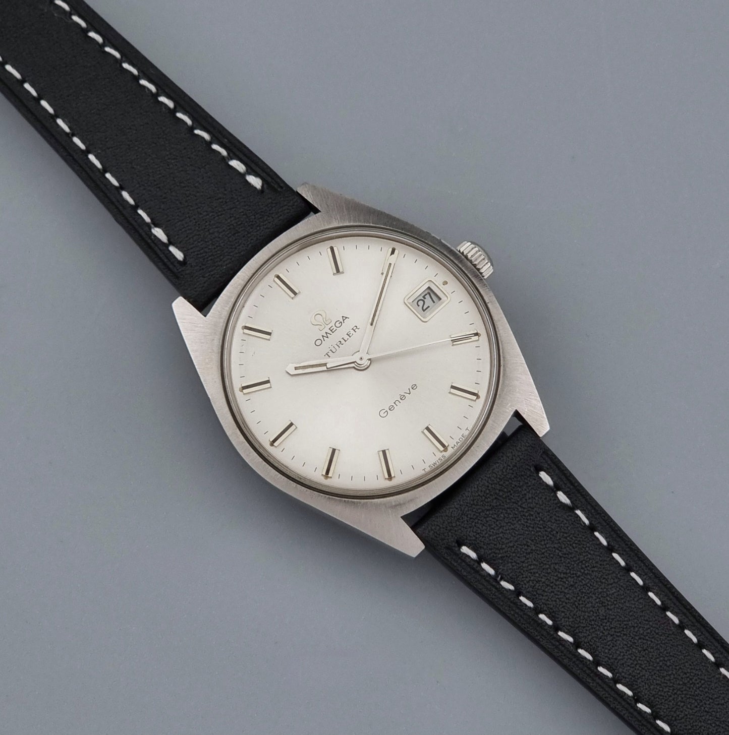 Vintage Omega watch Geneve 136.041 Co-signed Türler with a stainless steel case,  and a black leather strap with white stitching, shown on a gray background dial