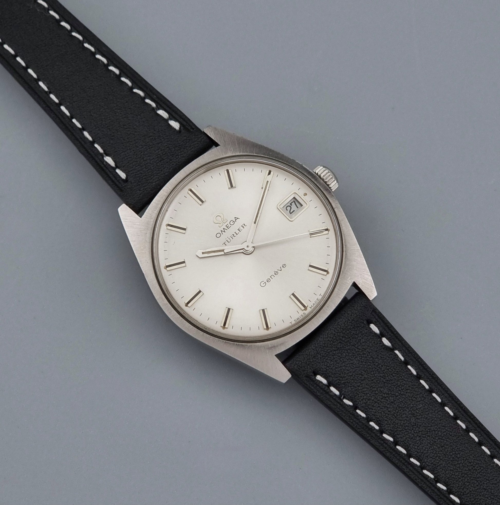 Vintage Omega watch Geneve 136.041 Co-signed Türler with a stainless steel case,  and a black leather strap with white stitching, shown on a gray background dial
