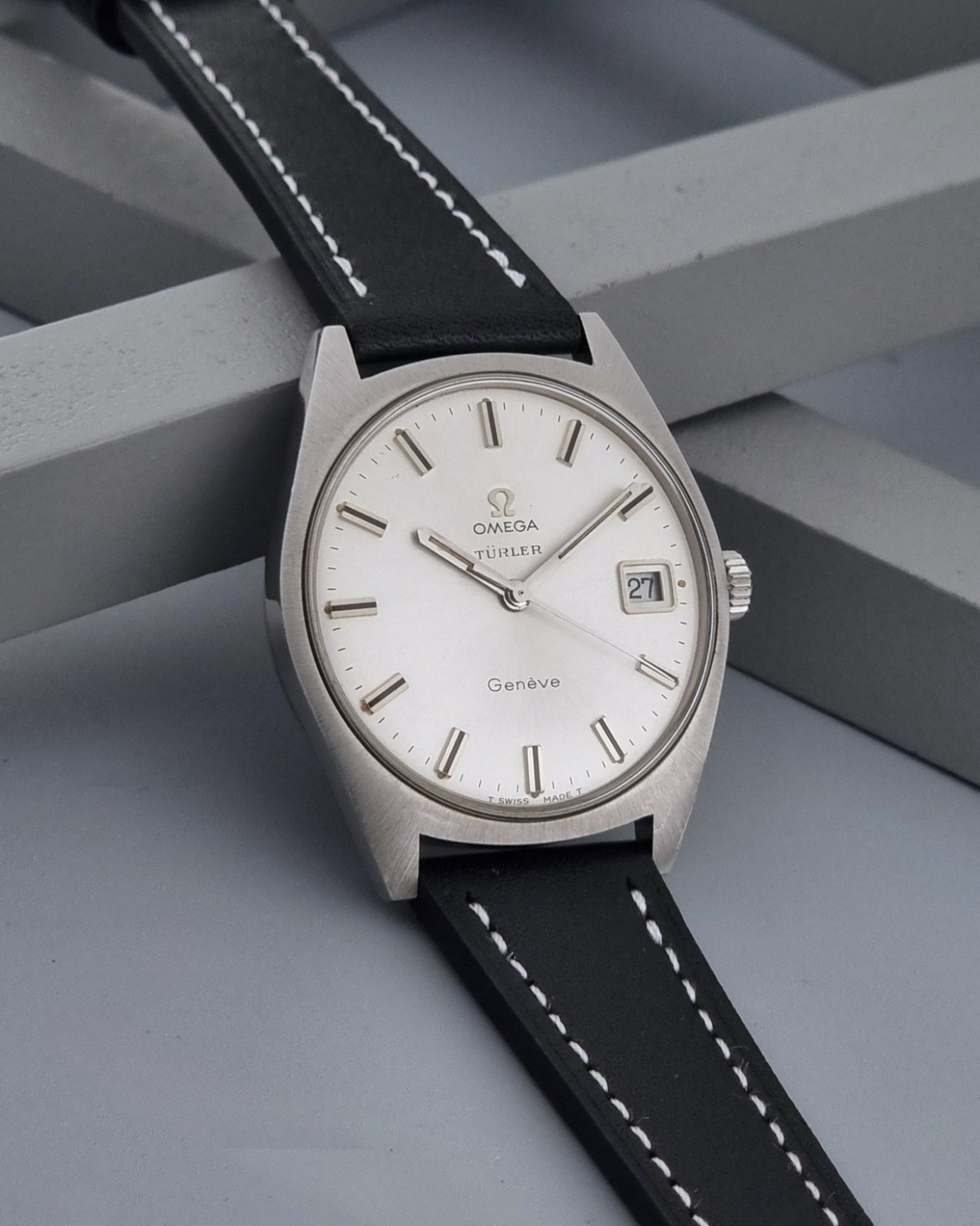 Vintage Omega watch Geneve 136.041 Co-signed Türler with a stainless steel case,  and a black leather strap with white stitching, shown on a gray background side view