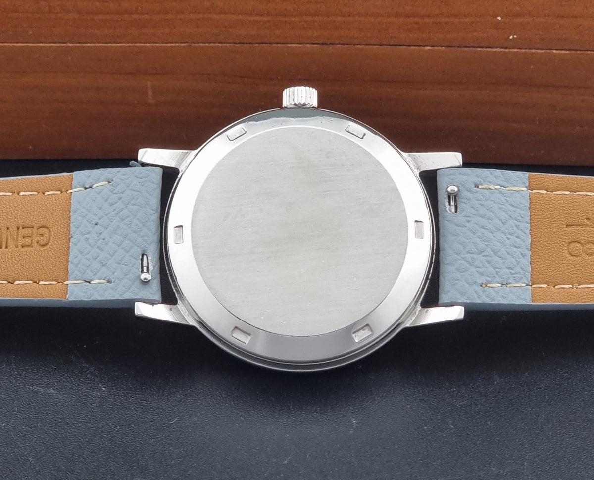 Close-up of a wristwatch from the back, showing a brushed silver-tone case and a grey leather strap with tan end sections on a dark surface.