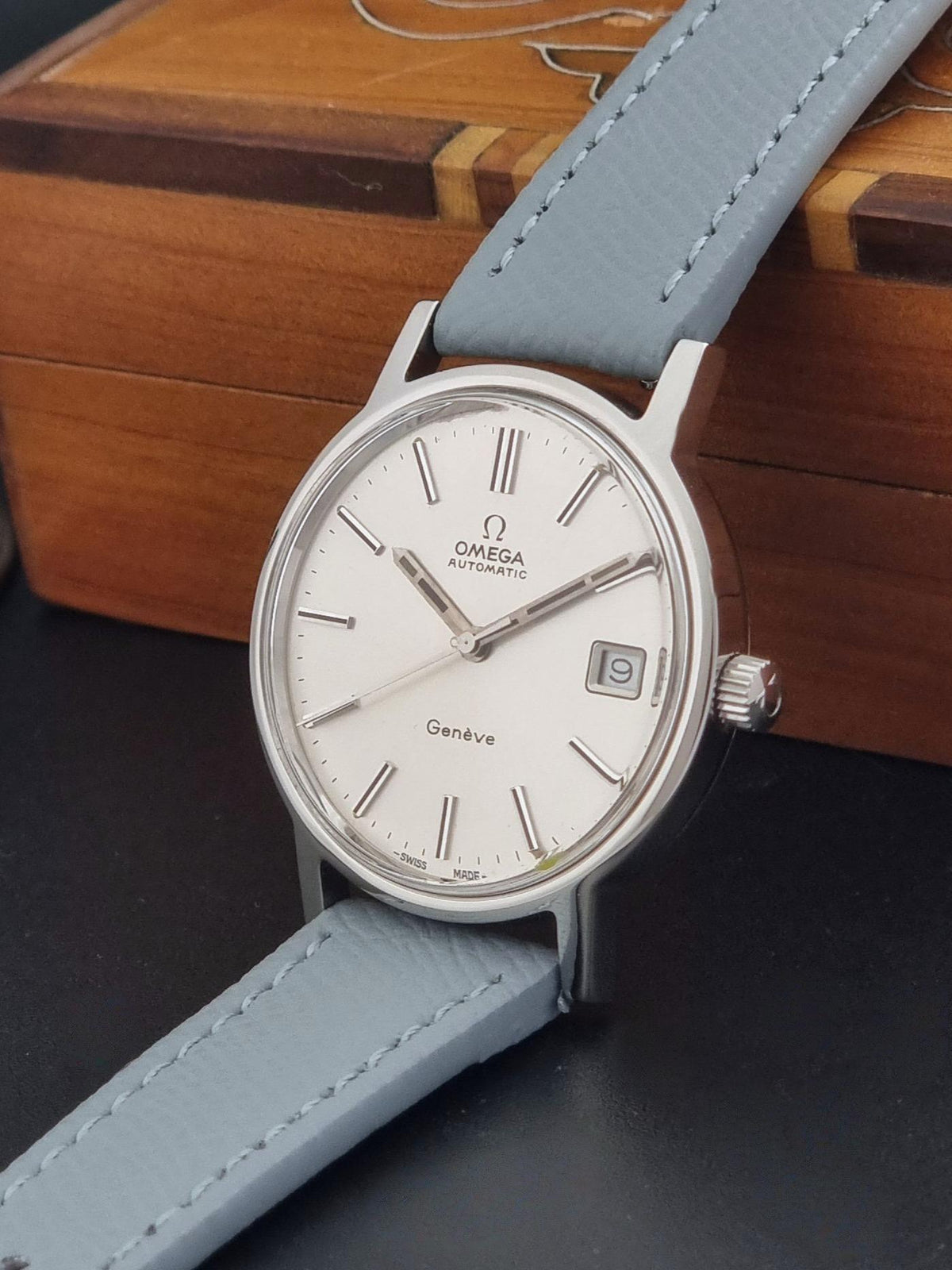 Vintage Omega Geneve 166.0163 Wristwatch with a silver-toned case, white dial, and light grey leather strap, resting on a dark surface with a wooden box in the background.