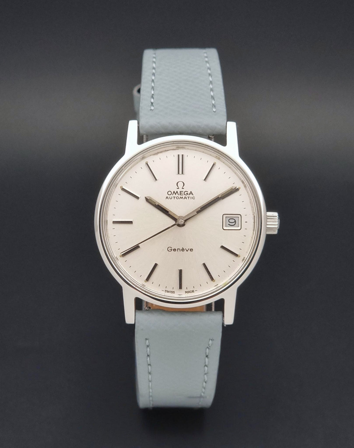 Vintage Omega Geneve 166.0163 Wristwatch with a silver-tone case, white dial, and light grey leather strap, shown against a dark gray background.