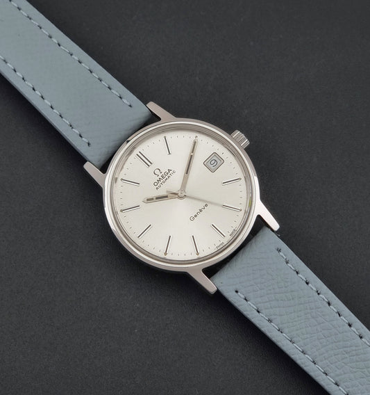Vintage Omega Geneve 166.0163 Wristwatch with a light grey leather strap and a silver-toned stainless steel case, displayed diagonally on a dark gray textured background.