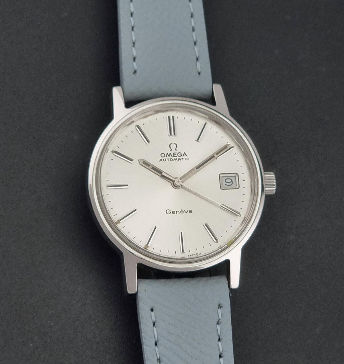 Vintage Omega Geneve 166.0163 Wristwatch with a silver-toned case, white dial, and a light grey leather strap, shown against a dark gray background.