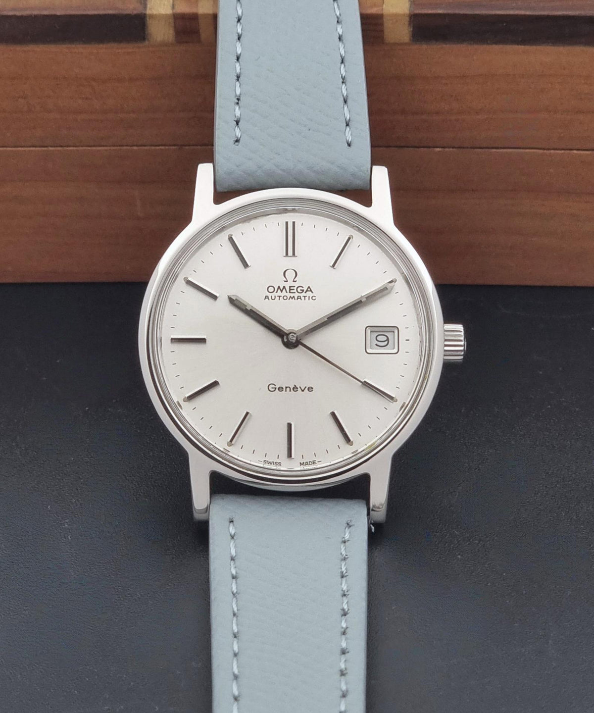 Vintage Omega Geneve 166.0163 Wristwatch with a silver-toned round case, white dial, and light grey leather strap, displayed against a dark surface with a wooden background.
