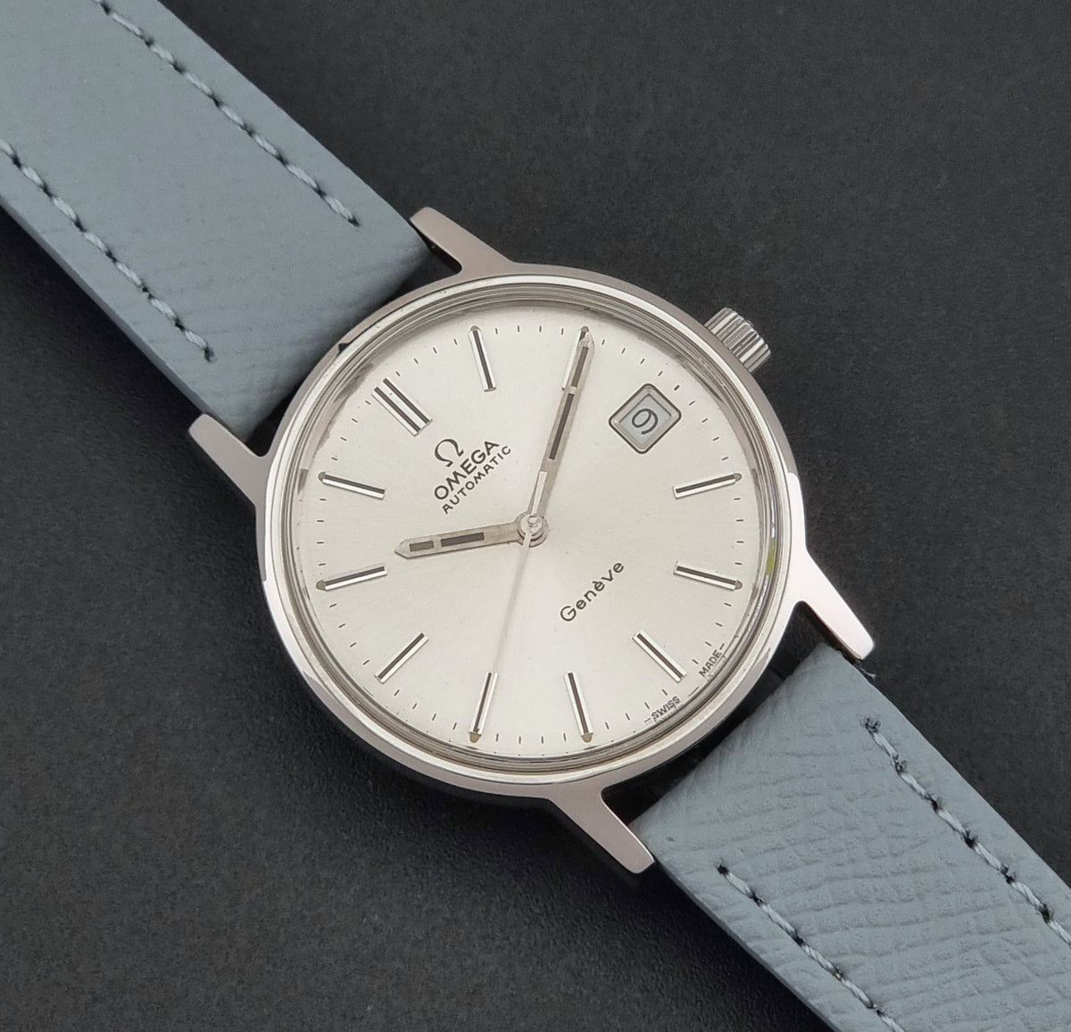 Vintage Omega Geneve 166.0163 Wristwatch with a silver-toned case, white dial, and light grey leather strap, shown diagonally on a dark textured background.