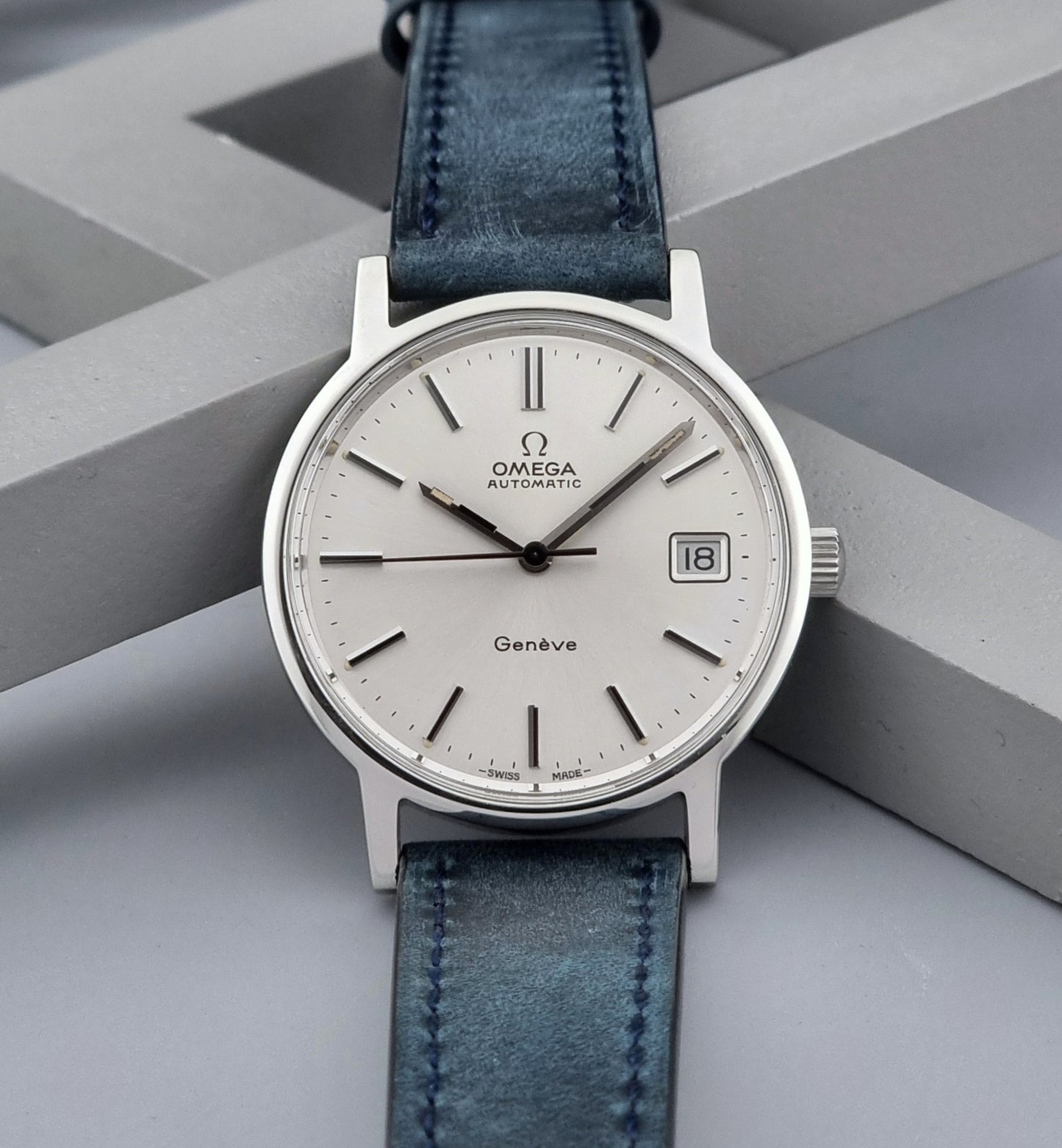 Vintage Omega Geneve 166.0163 wristwatch with blue leather strap on a geometric gray background