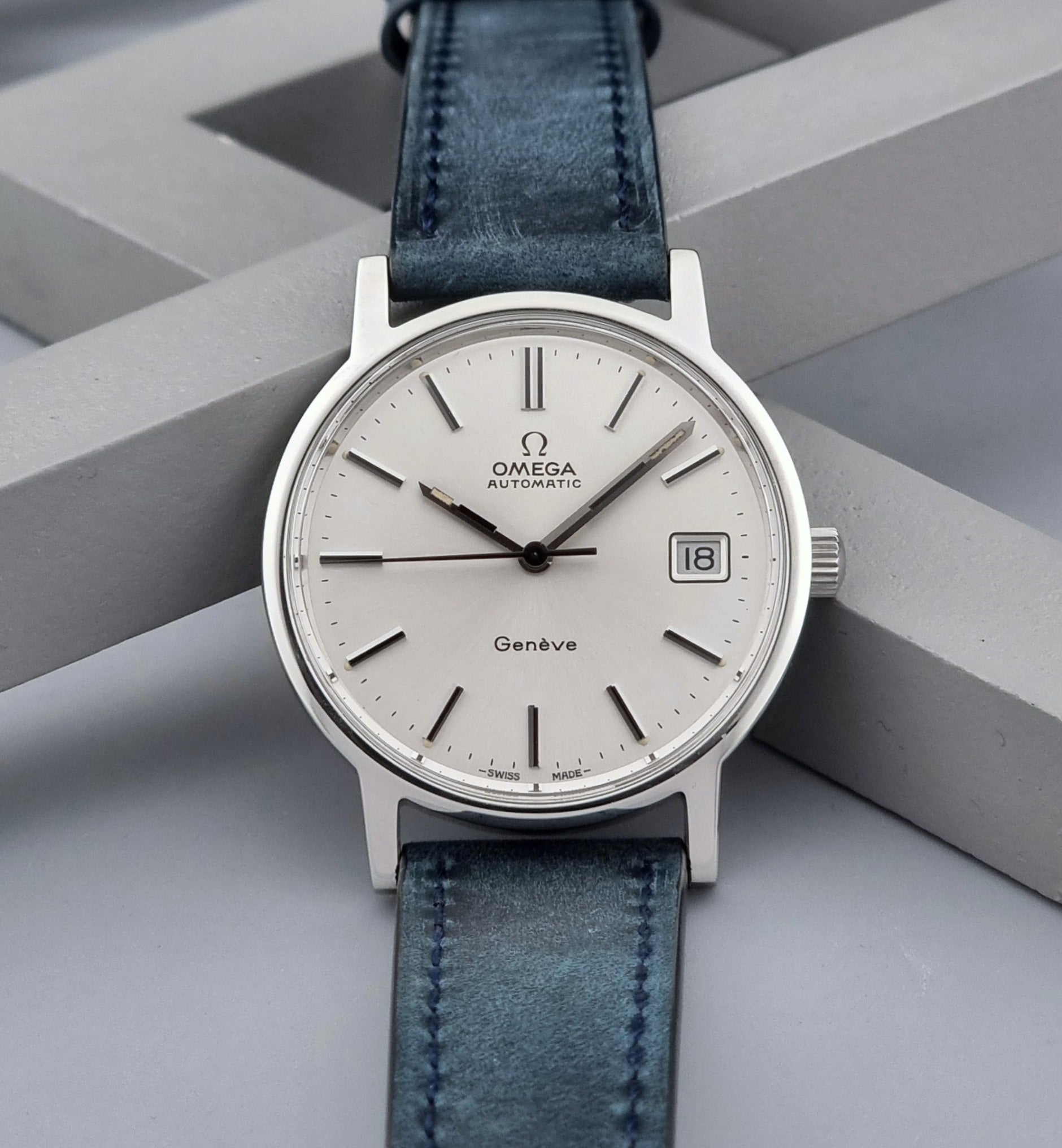 Vintage Omega Geneve 166.0163 wristwatch with blue leather strap on a geometric gray background