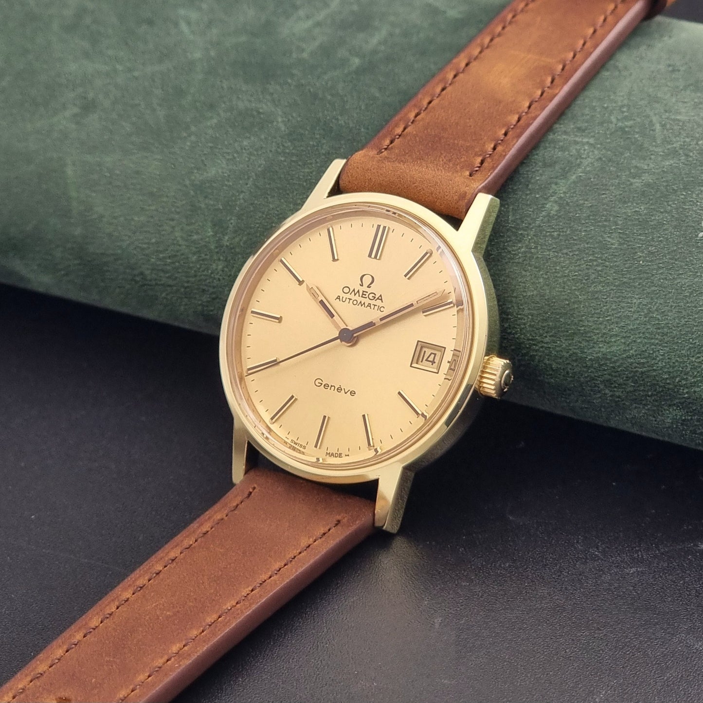 Vintage Omega Geneve Wristwatch 166.0163 in gold with brown leather strap on a dark gray background crown view