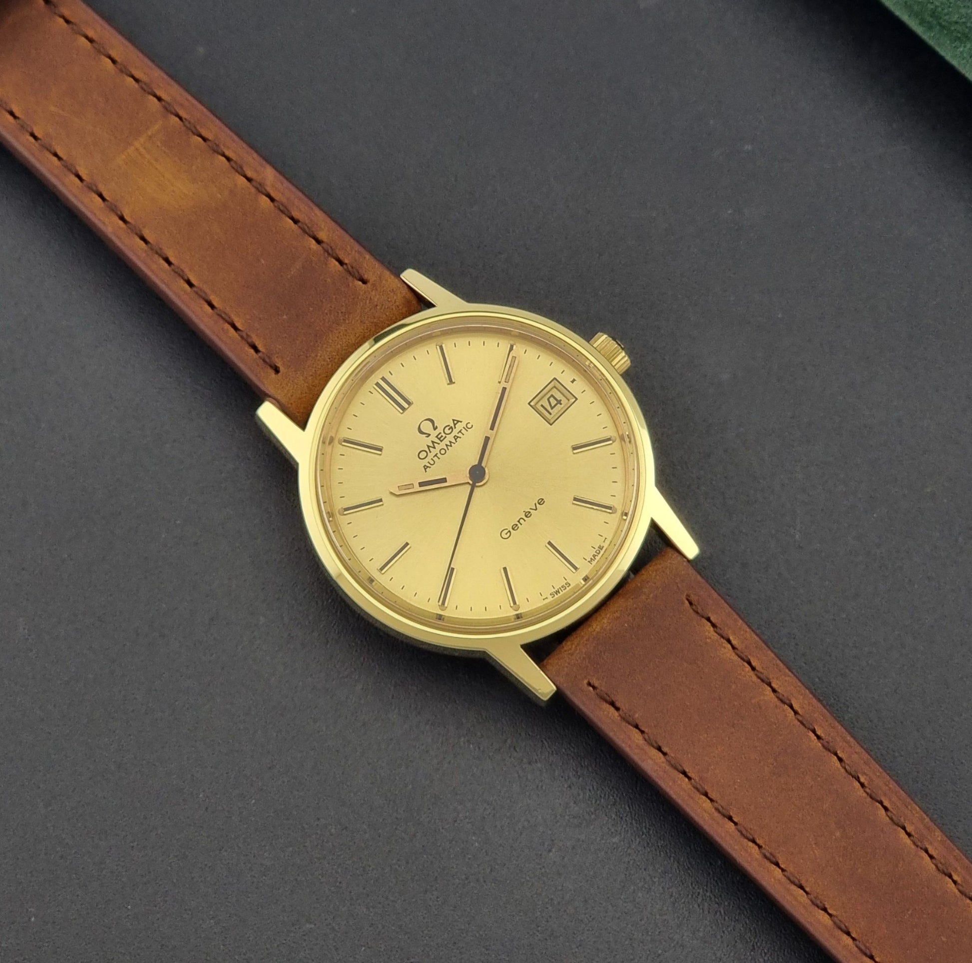 Vintage Omega Geneve Wristwatch 166.0163 in gold with brown leather strap on a dark gray background dial