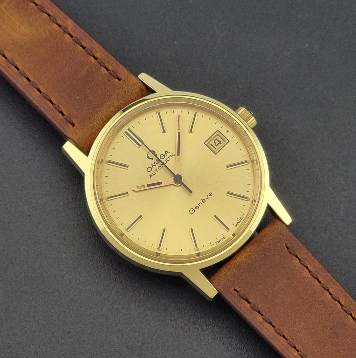 Vintage Omega Geneve Wristwatch 166.0163 in gold with brown leather strap on a dark gray background close up dial view