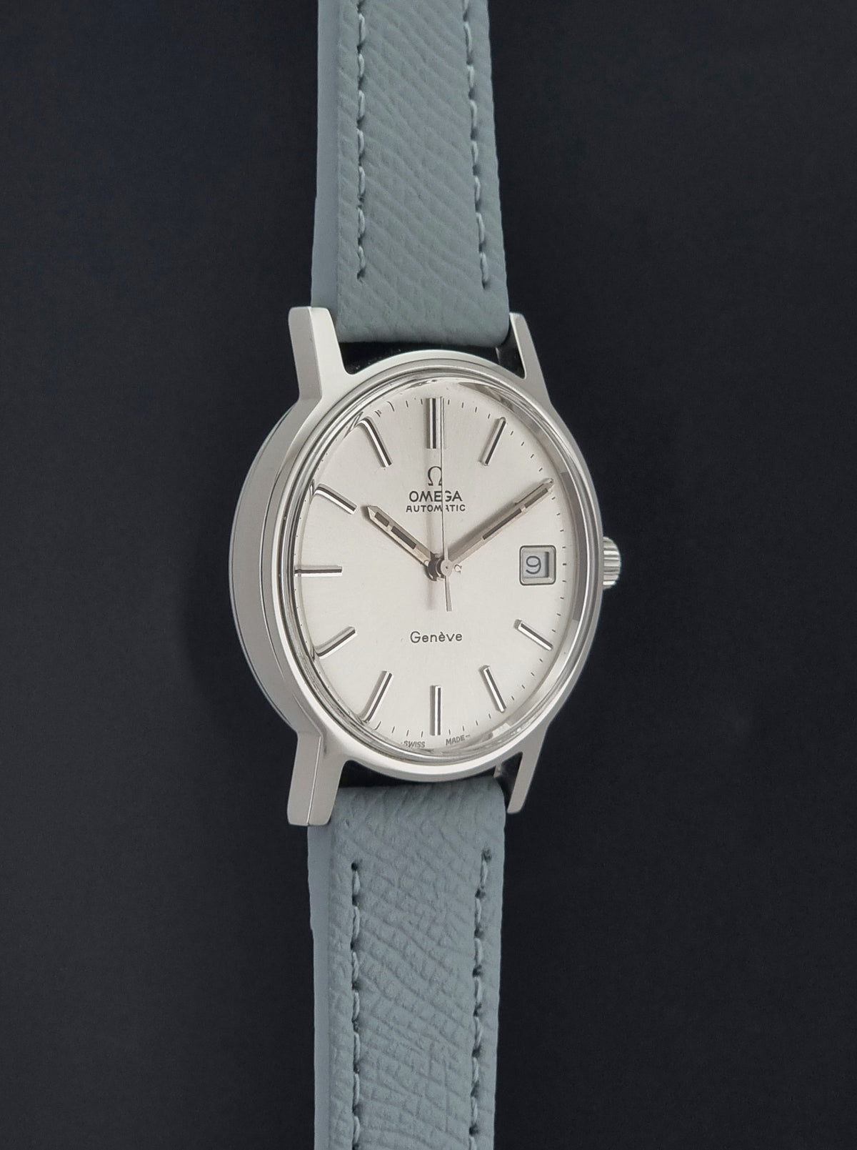 Vintage Omega Geneve 166.0163 Wristwatch with a silver-toned case, white dial, and a light grey textured leather strap, shown against a dark background.