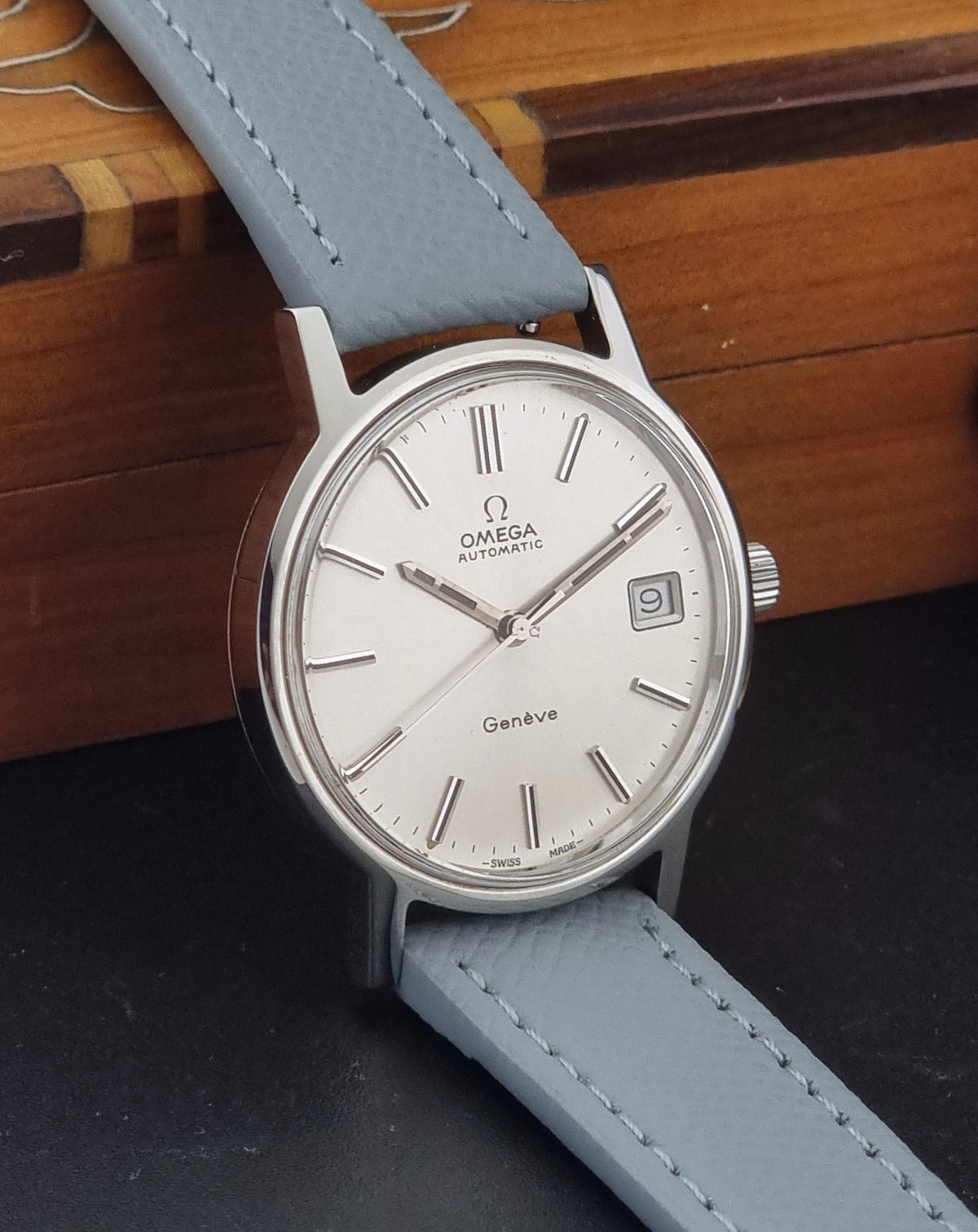 Vintage Omega Geneve 166.0163 Wristwatch with a silver stainless steel case, white dial, and a light gray leather strap, resting on a dark surface with a wooden box in the background.