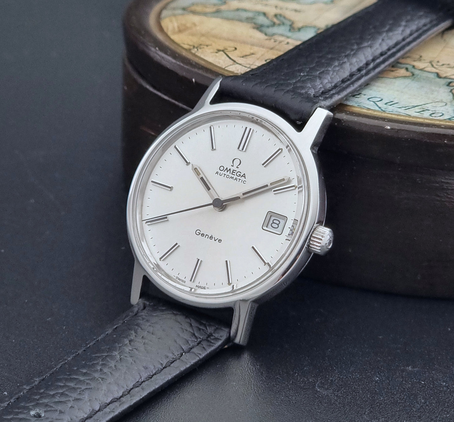 Vintage Omega Geneve Wristwatch 166.0163 with black leather strap and silver case on a dark gray background crown view