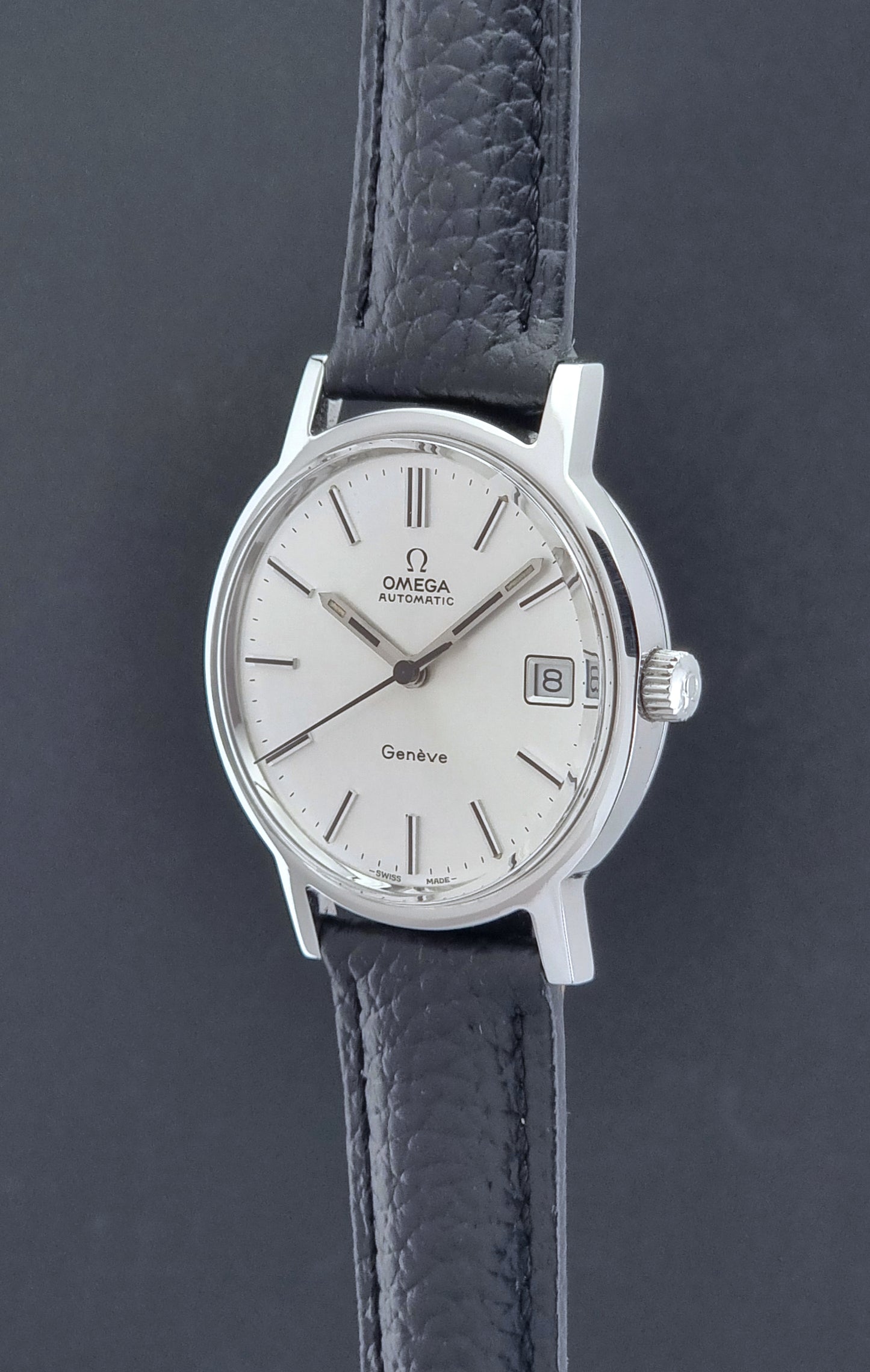 Vintage Omega Geneve Wristwatch 166.0163 with black leather strap and silver case on a dark gray background crown profile view