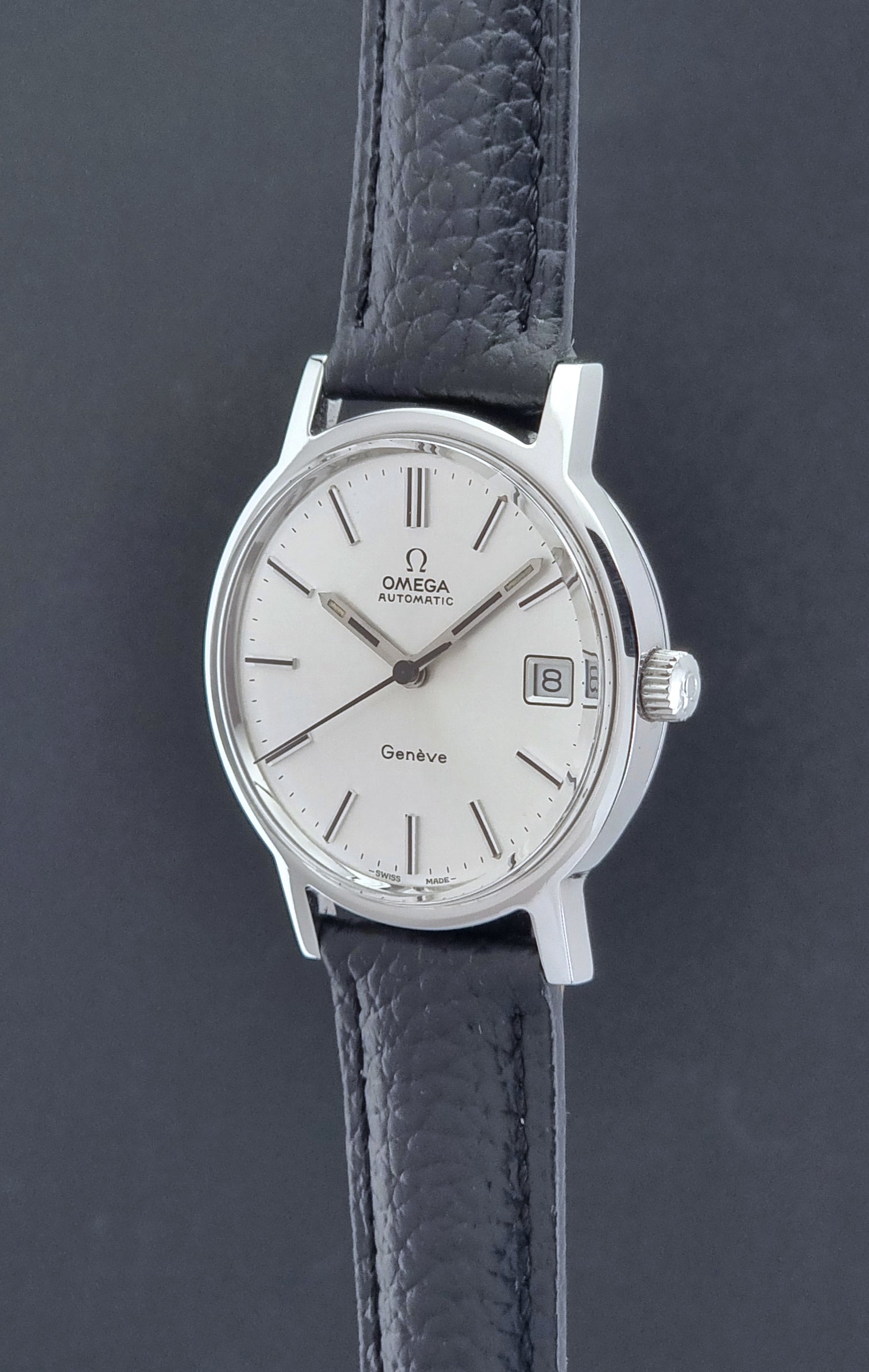 Vintage Omega Geneve Wristwatch 166.0163 with black leather strap and silver case on a dark gray background crown profile view