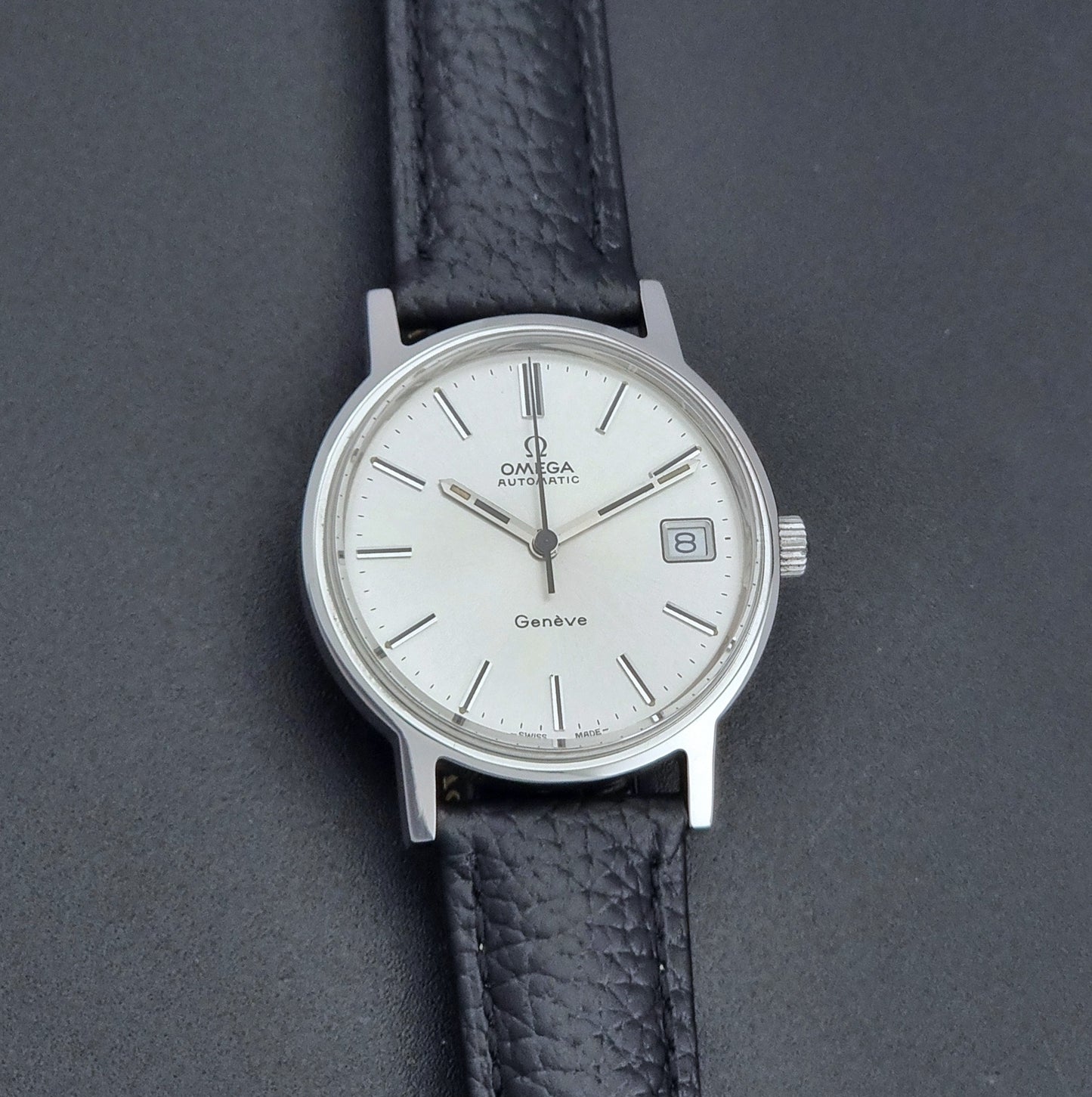 Vintage Omega Geneve Wristwatch 166.0163 with black leather strap and silver case on a dark gray background front view