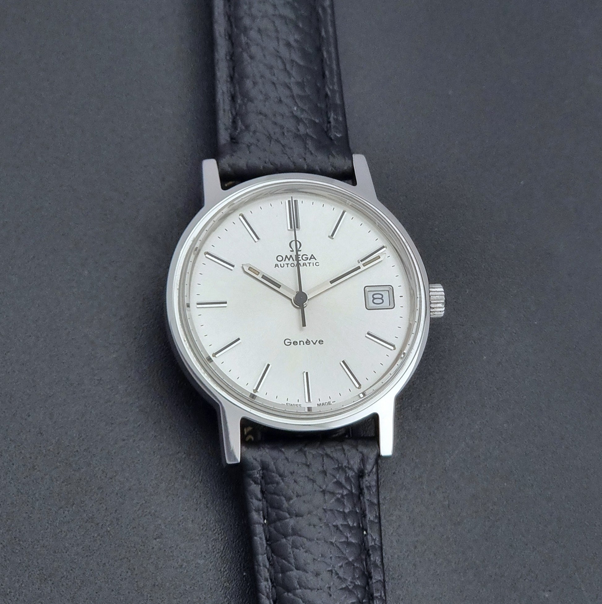 Vintage Omega Geneve Wristwatch 166.0163 with black leather strap and silver case on a dark gray background front view