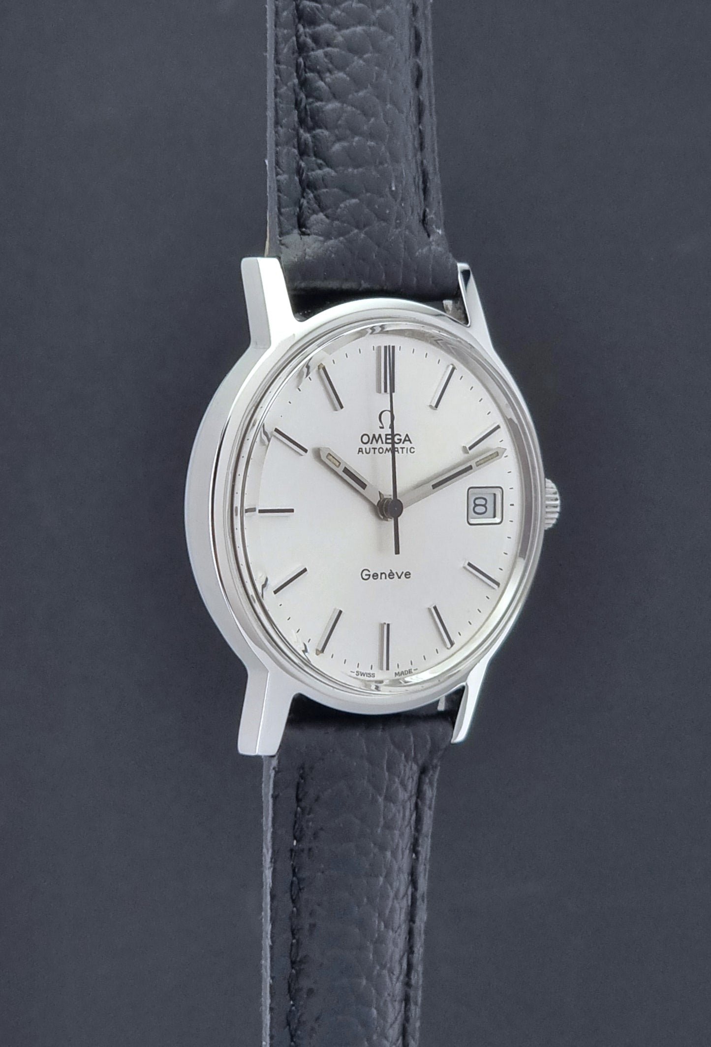 Vintage Omega Geneve Wristwatch 166.0163 with black leather strap and silver case on a dark gray background side profile view