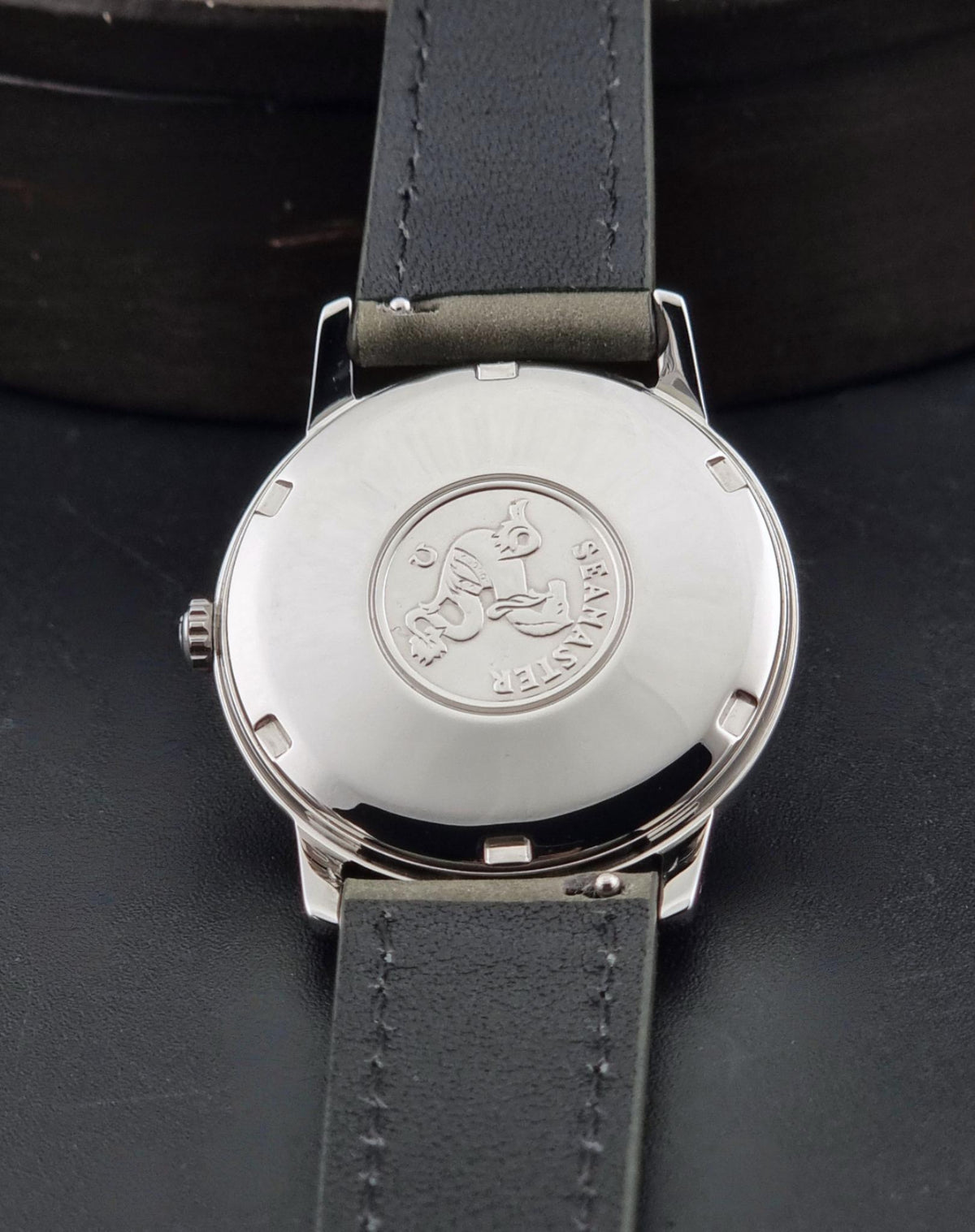 Close-up of the back of a wristwatch with a round silver-tone case and a dark gray leather strap, resting on a dark surface. Engraved Seamaster