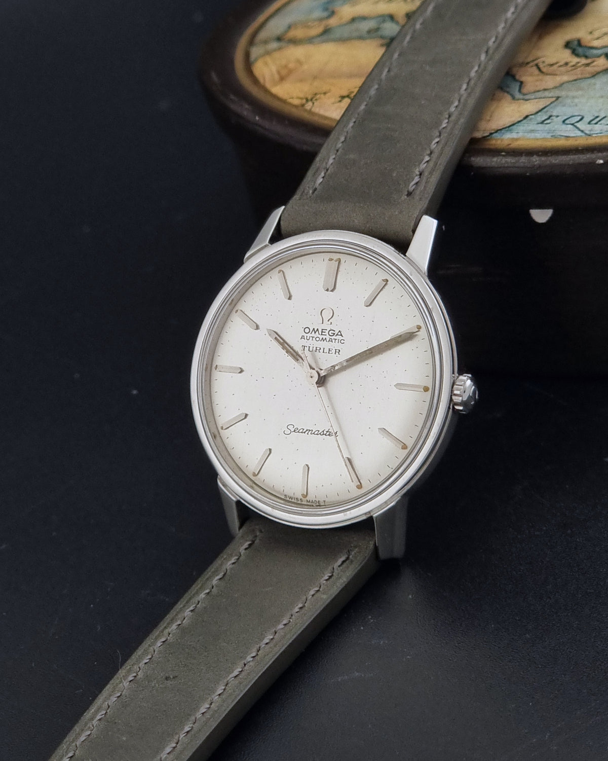 Vintage Omega Wristwatch Seamaster Turler 165.002 with a silver-toned case, white dial, and grey leather strap, displayed diagonally on a dark surface with a vintage map in the background.