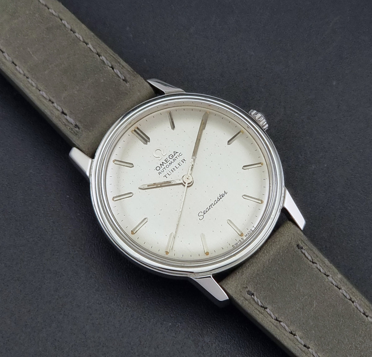 An Omega Seamaster watch with a silver-toned stainless steel case, cream-colored dial, and an grey leather strap, shown on a dark textured background.