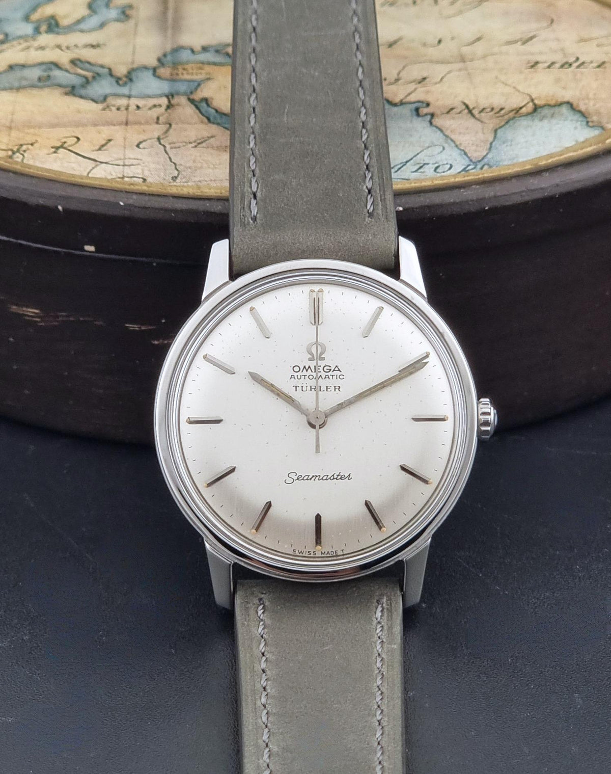 An Omega Seamaster Automatic watch with a silver-toned stainless steel case, white dial, and grey leather strap, displayed in front of a vintage world map and dark wooden box.