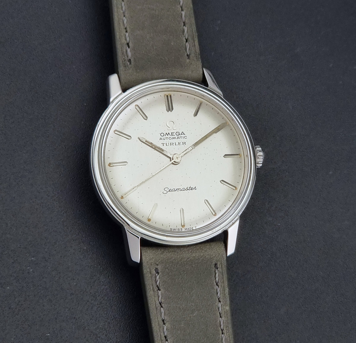 Vintage Omega Wristwatch Seamaster Turler 165.002 with a silver-toned stainless steel case, white dial, and grey leather strap, shown on a dark textured background.