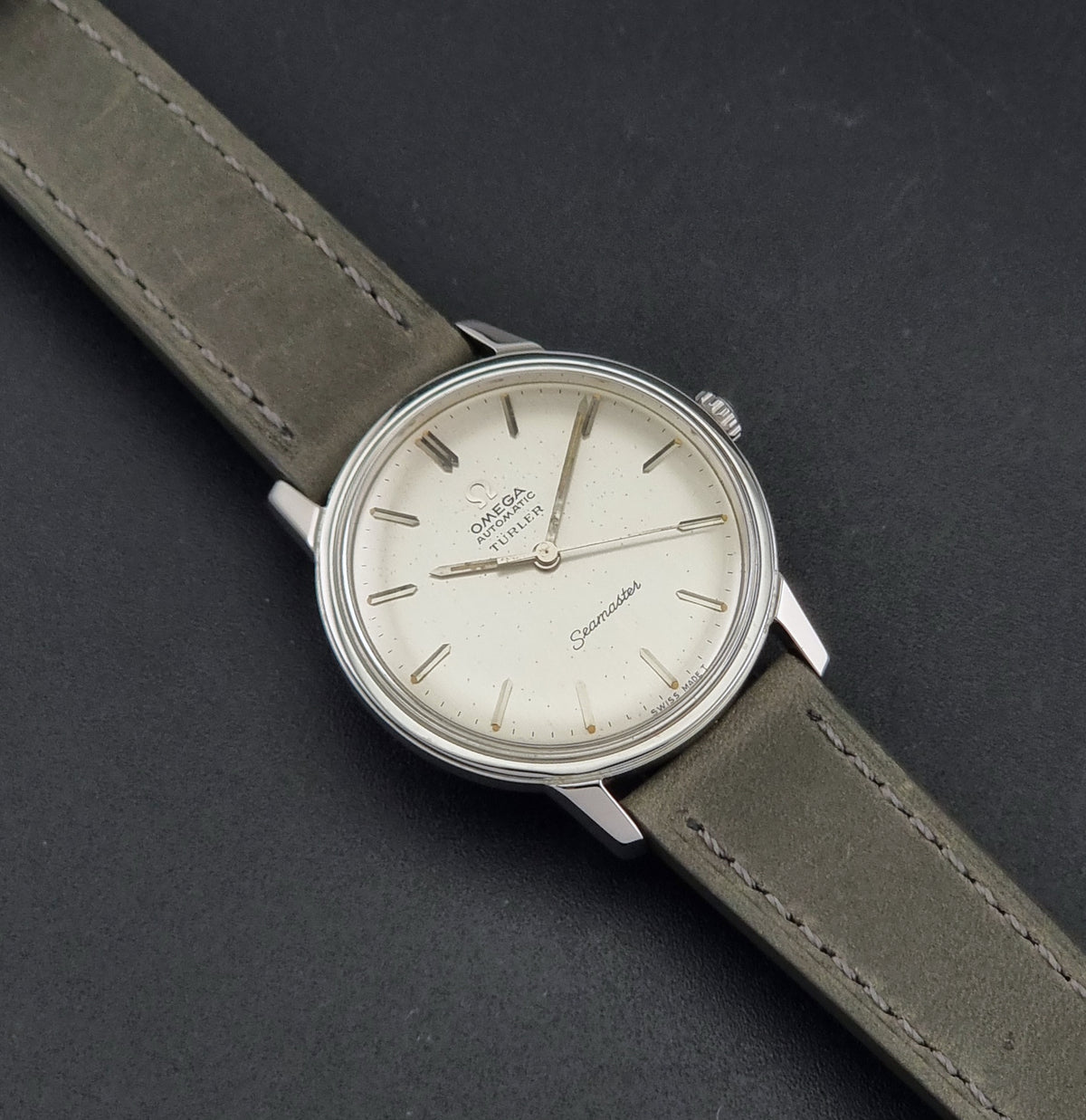 Vintage Omega Wristwatch Seamaster Turler 165.002 with a silver-toned case, cream-colored dial, and olive green leather strap, shown on a dark textured background.