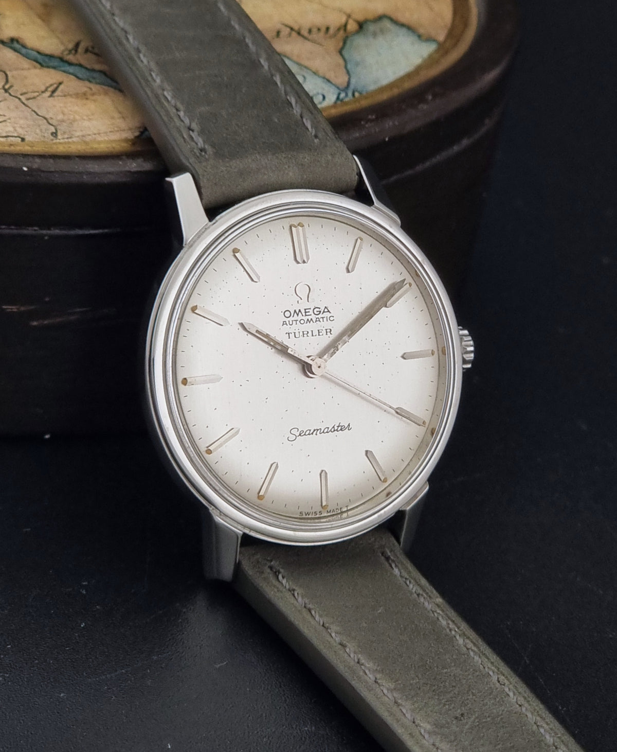 Omega Seamaster Automatic wristwatch with a silver-toned oval case, white dial, and grey leather strap, resting on a dark surface with a vintage map box in the background.
