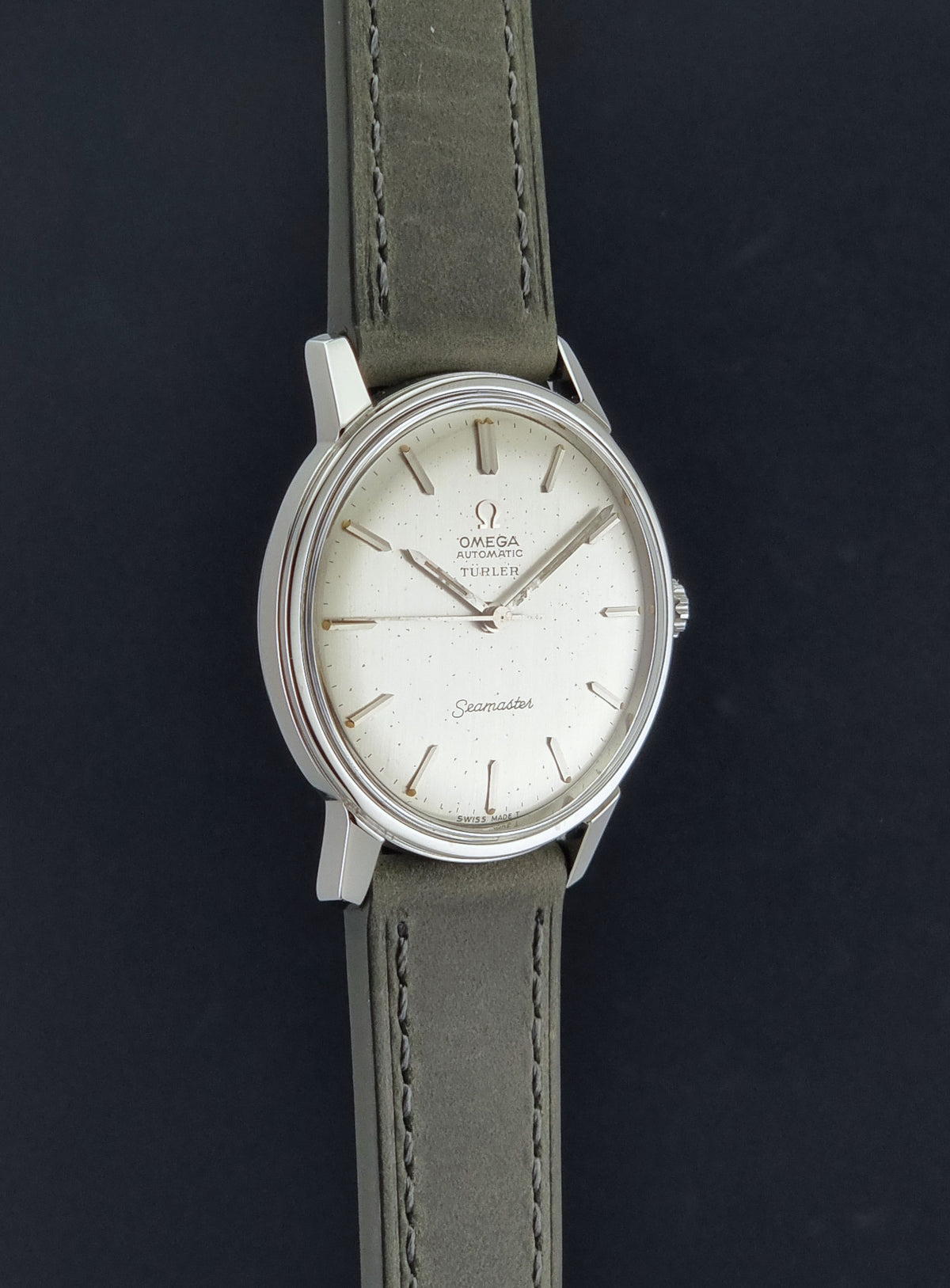Vintage Omega Wristwatch Seamaster Turler 165.002 with a silver-toned oval case, white dial, and grey leather strap, shown against a dark background.
