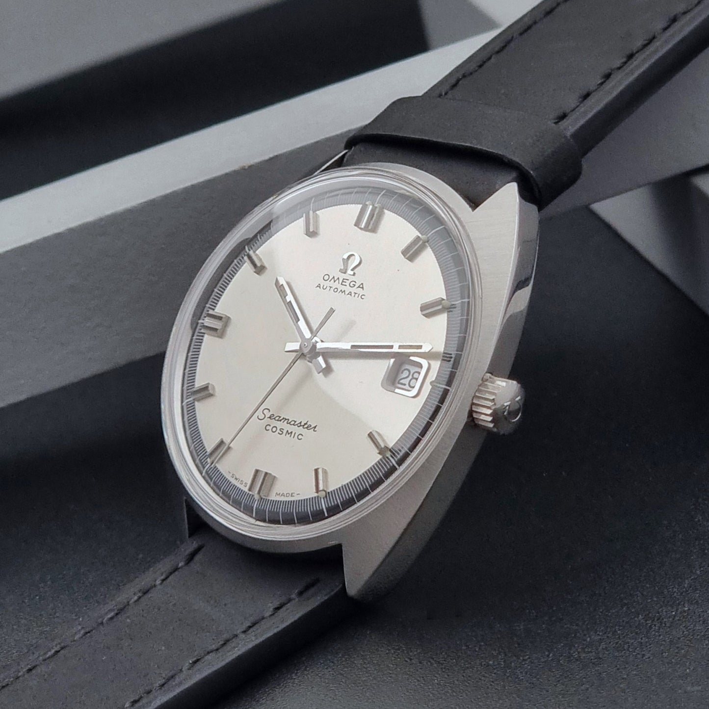 Vintage Omega Wristwatch Seamaster Cosmic 166.026 with silver dial and black leather strap on a gray background crown side view