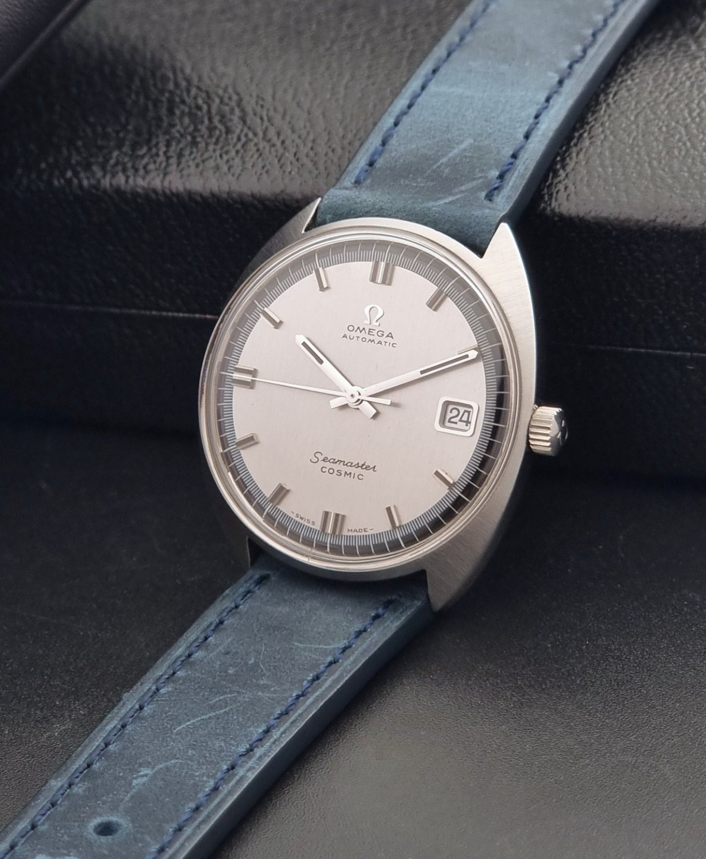 Vintage Omega 166.026 Seamaster Cosmic Wristwatch with blue leather strap and silver case on a dark gray background crown view