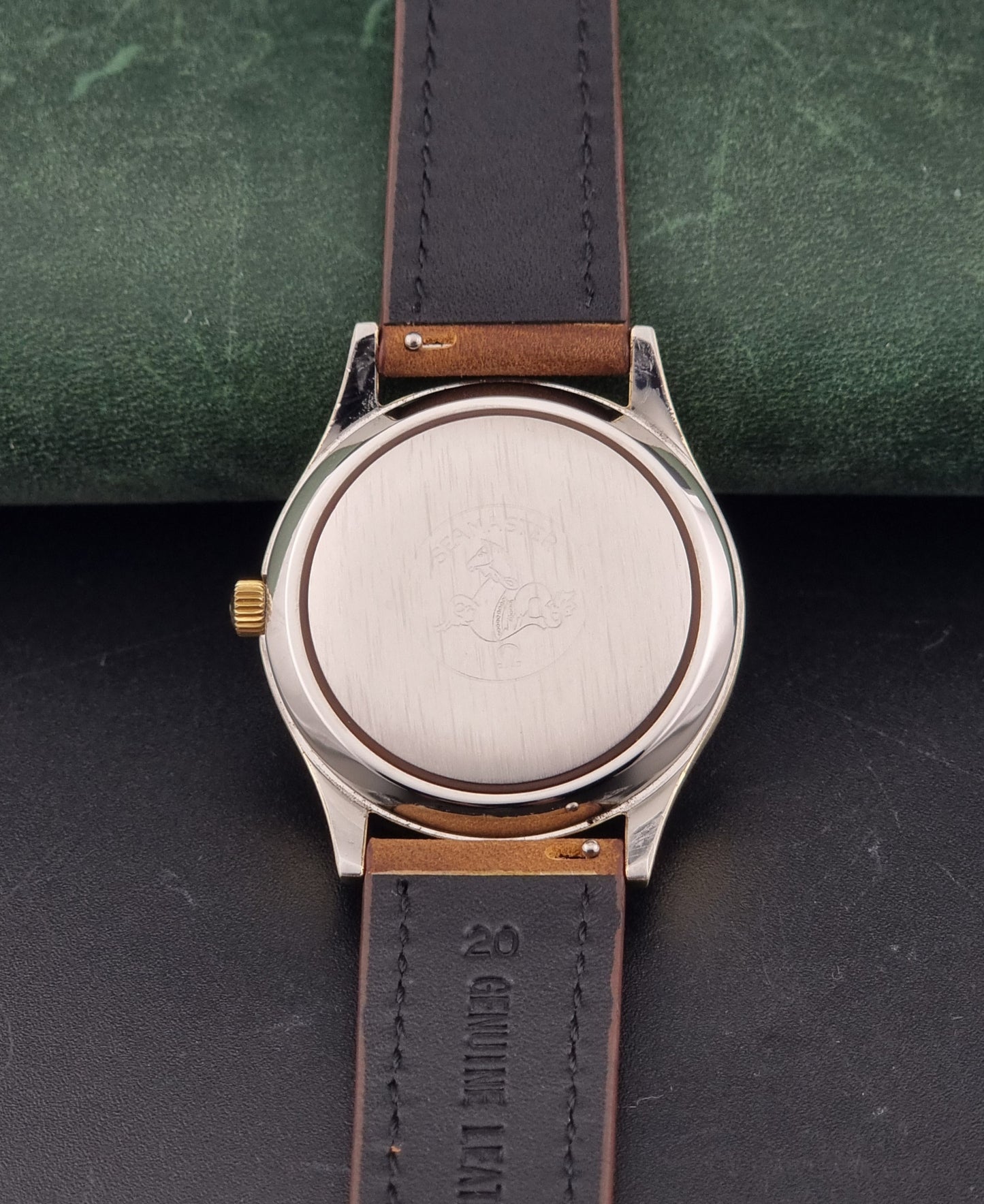 Close-up of the back of a Vintage Omega Wristwatch Seamaster Cosmic 2000 Jumbo 166.129 with a brushed silver-tone case, a gold-tone crown, and a brown leather strap on a dark textured background.