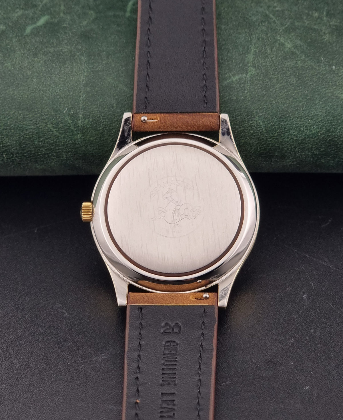 Close-up of the back of a Vintage Omega Wristwatch Seamaster Cosmic 2000 Jumbo 166.129 with a brushed silver-tone case, a gold-tone crown, and a brown leather strap on a dark textured background.