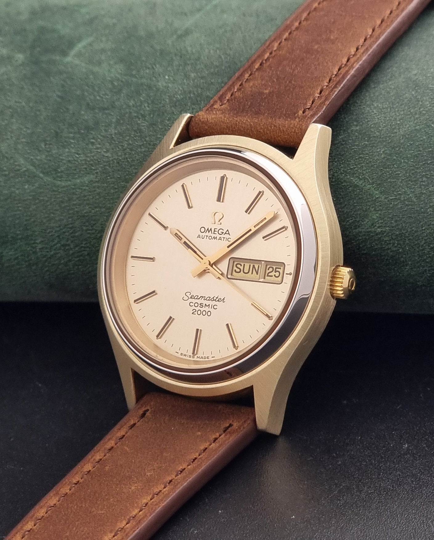 An Vintage Omega Wristwatch Seamaster Cosmic 2000 Jumbo 166.129 with a gold-tone case, gold dial, and brown leather strap, resting on a dark textured surface with a green background.