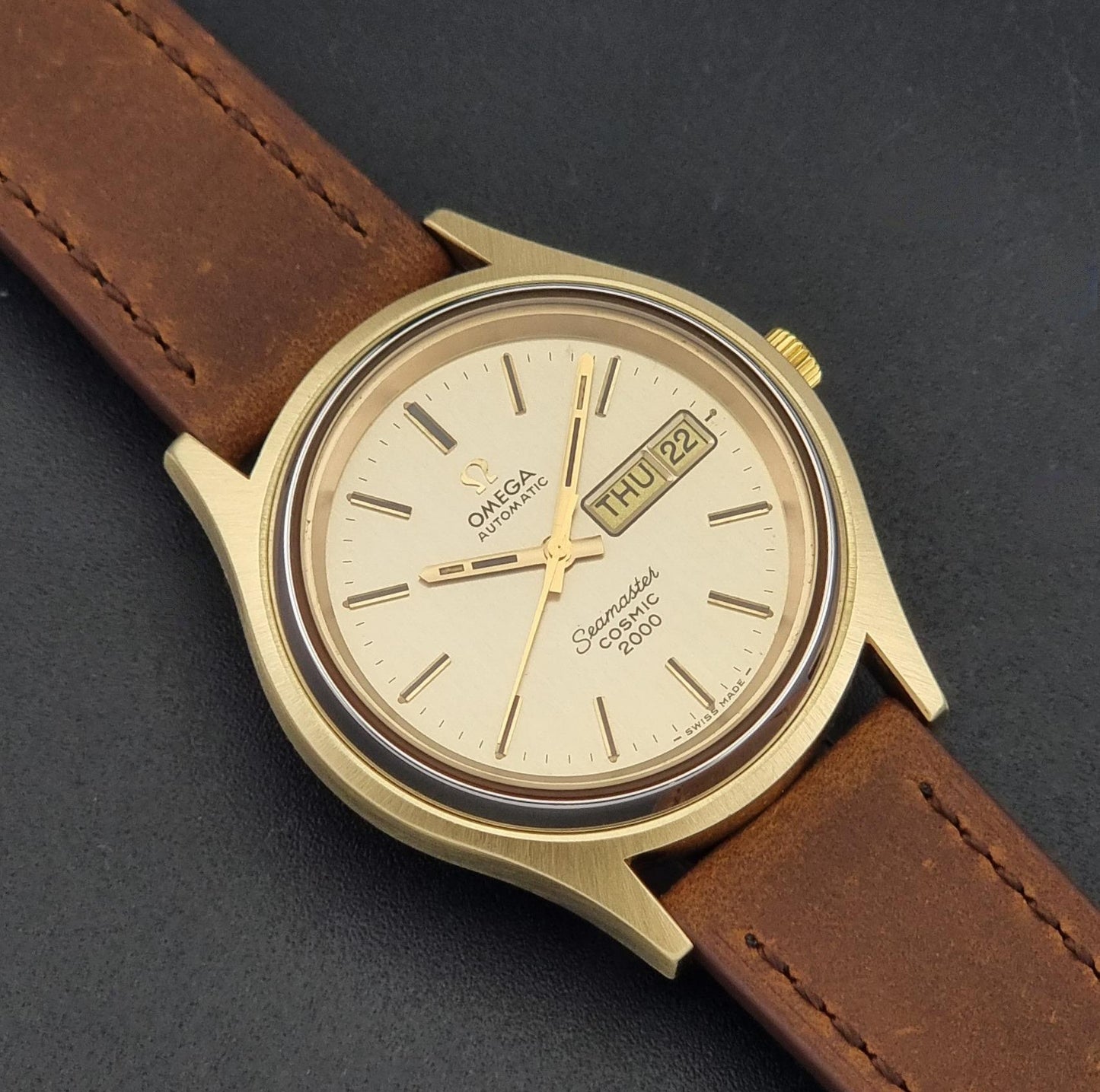 A Vintage Omega Wristwatch Seamaster Cosmic 2000 Jumbo 166.129 with a gold-tone case, gold dial, and brown leather strap, shown on a dark textured background.