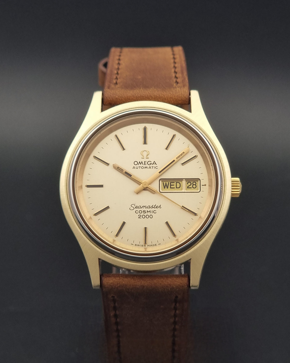 An Vintage Omega Wristwatch Seamaster Cosmic 2000 Jumbo 166.129 with a gold-tone case, gold dial, and a brown leather strap, shown against a dark gray background.