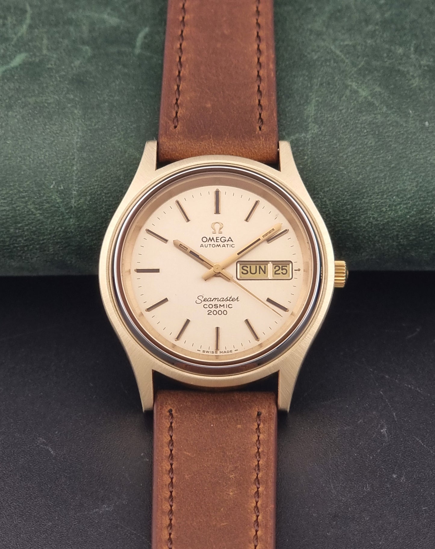 A Vintage Omega Wristwatch Seamaster Cosmic 2000 Jumbo 166.129 wristwatch with a gold-toned case, gold dial, and brown leather strap, displayed on a dark textured surface with a green background.